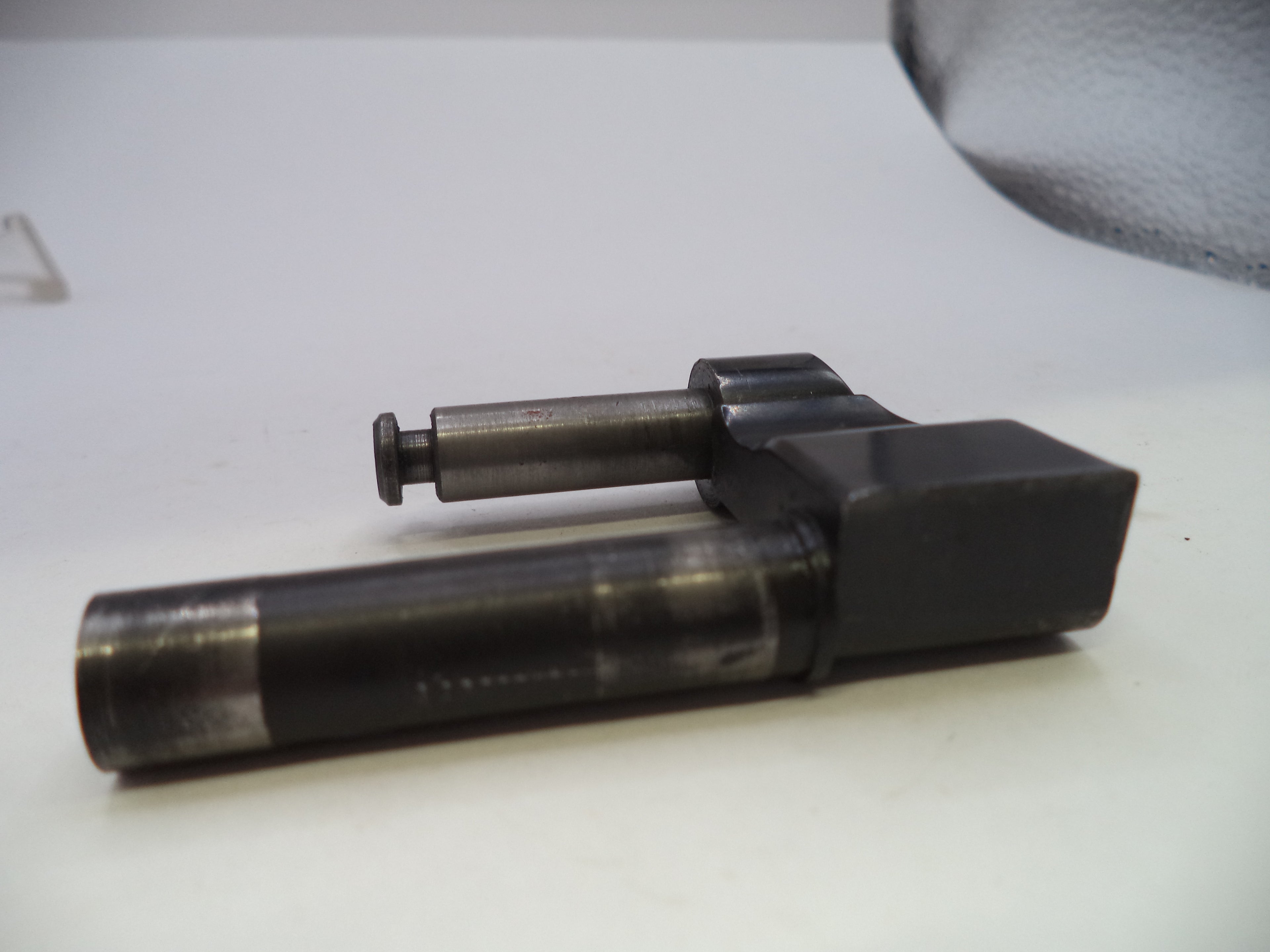 A used black and dark gray metal mechanical part, likely a pump or injector component, resting on a white surface.