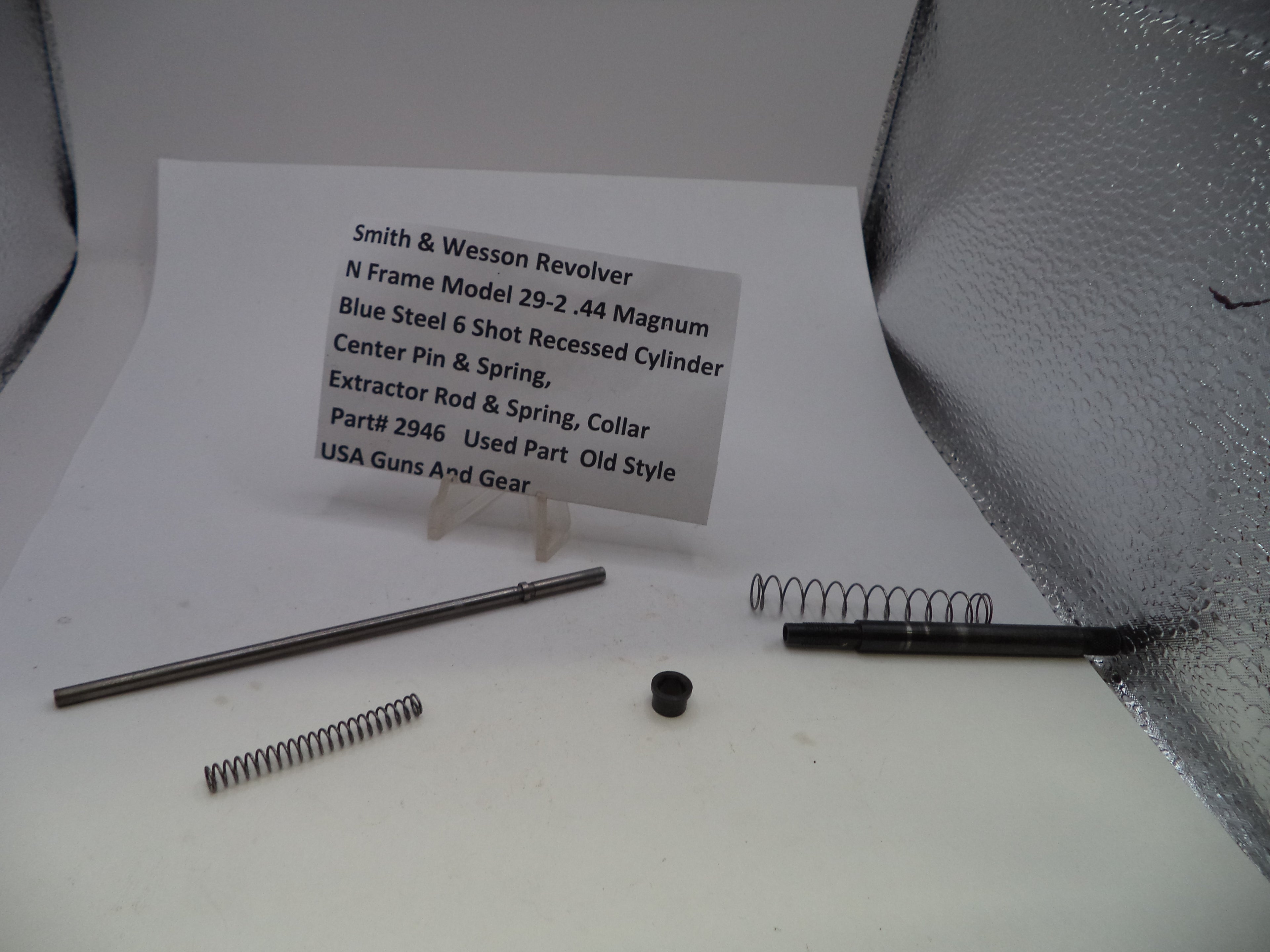 Smith & Wesson revolver replacement parts laid out on a white surface, including a long metal rod, two coiled springs, a small black cap, and a handwritten label card with part details.