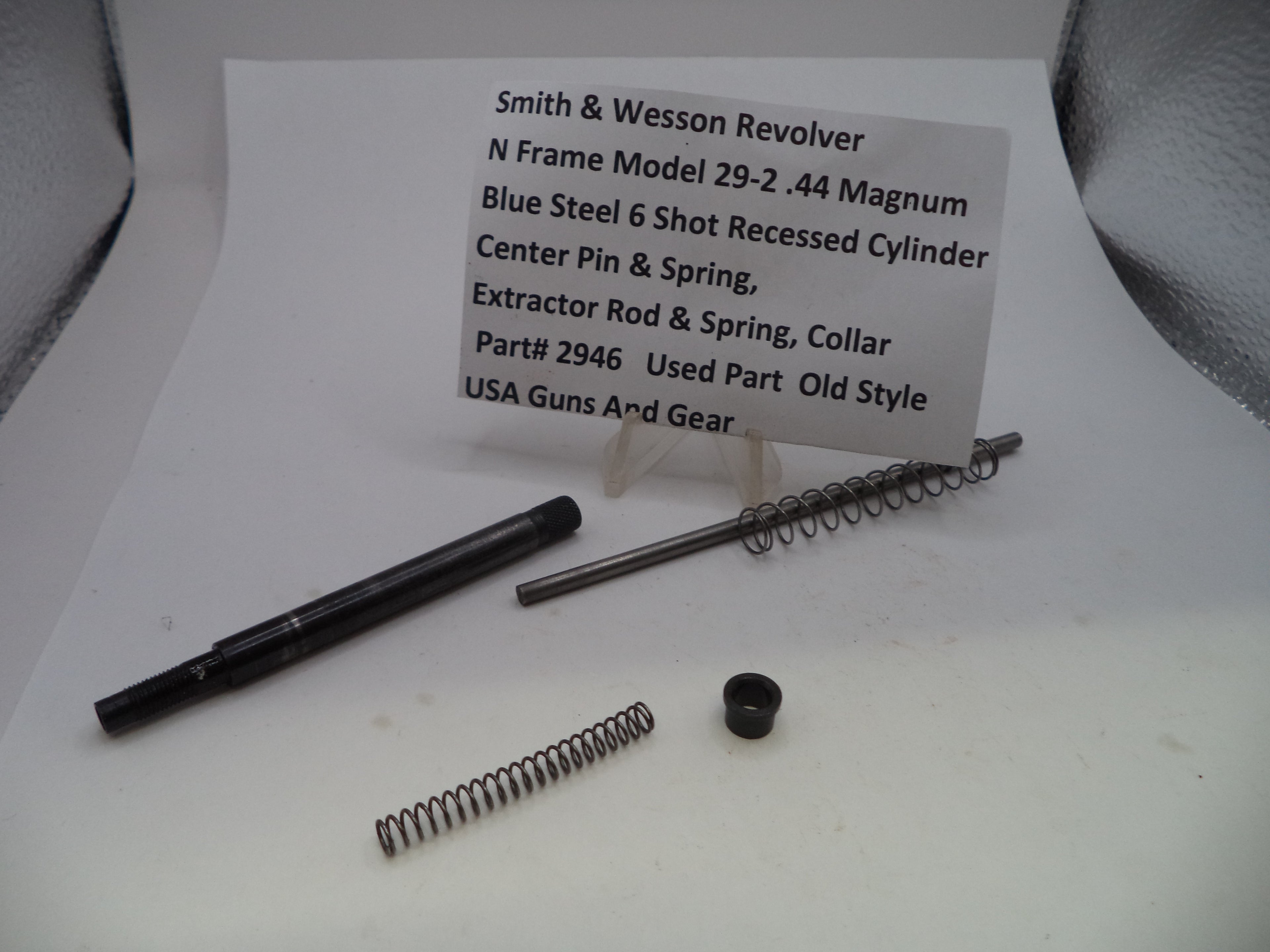 A used metal component set for a Smith & Wesson revolver, including an extractor pin and spring, a center pin and spring, and a collar, displayed on a white surface with a paper label card.