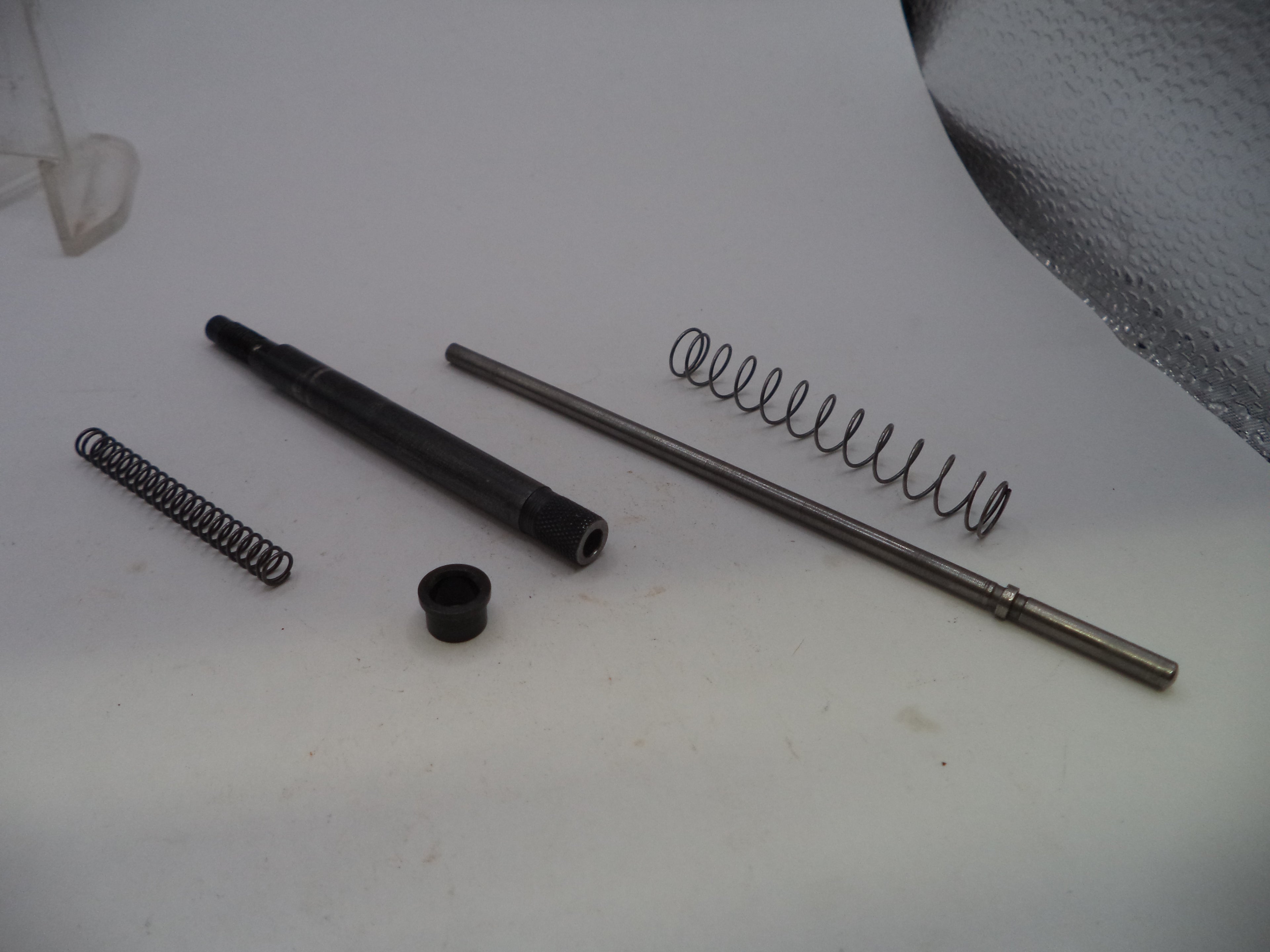 A disassembled mechanical component set featuring a long metal rod, a black cylindrical part, a black cap, and two coiled metal springs on a white surface.