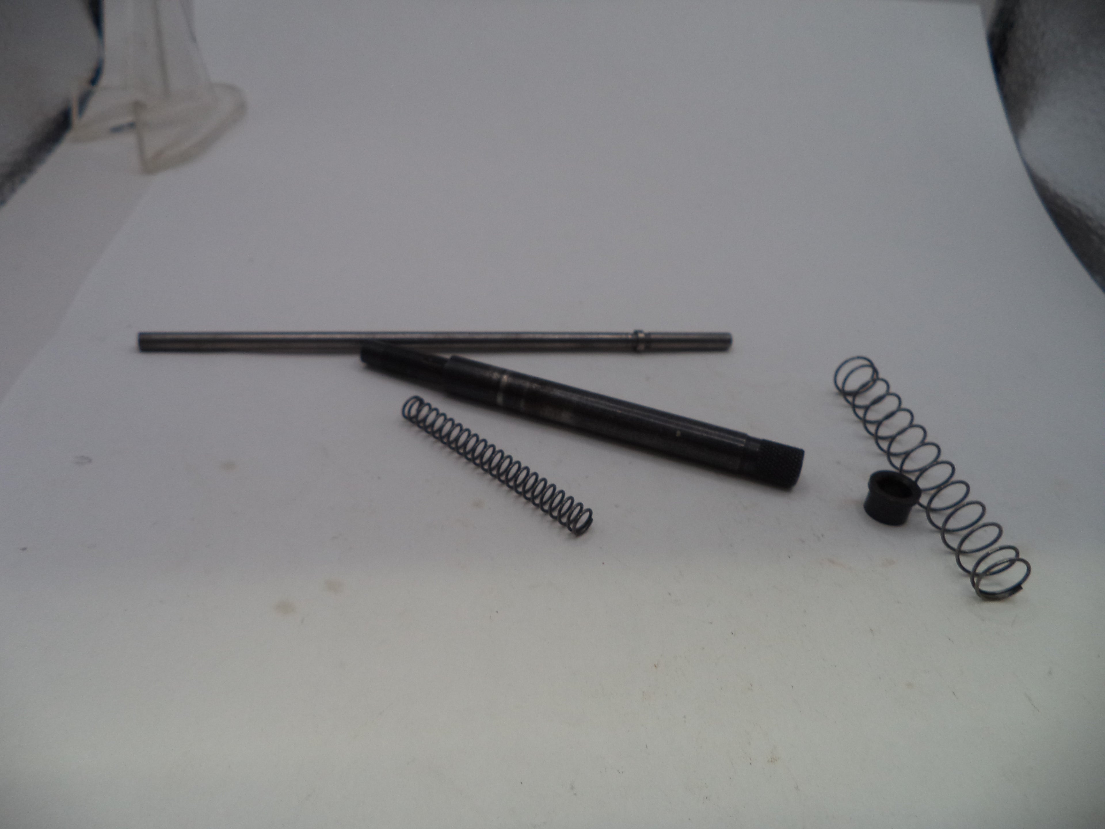 A set of metal mechanical parts, including a long rod, a black cylindrical component, two coiled springs, and a small black cap, arranged on a white surface.