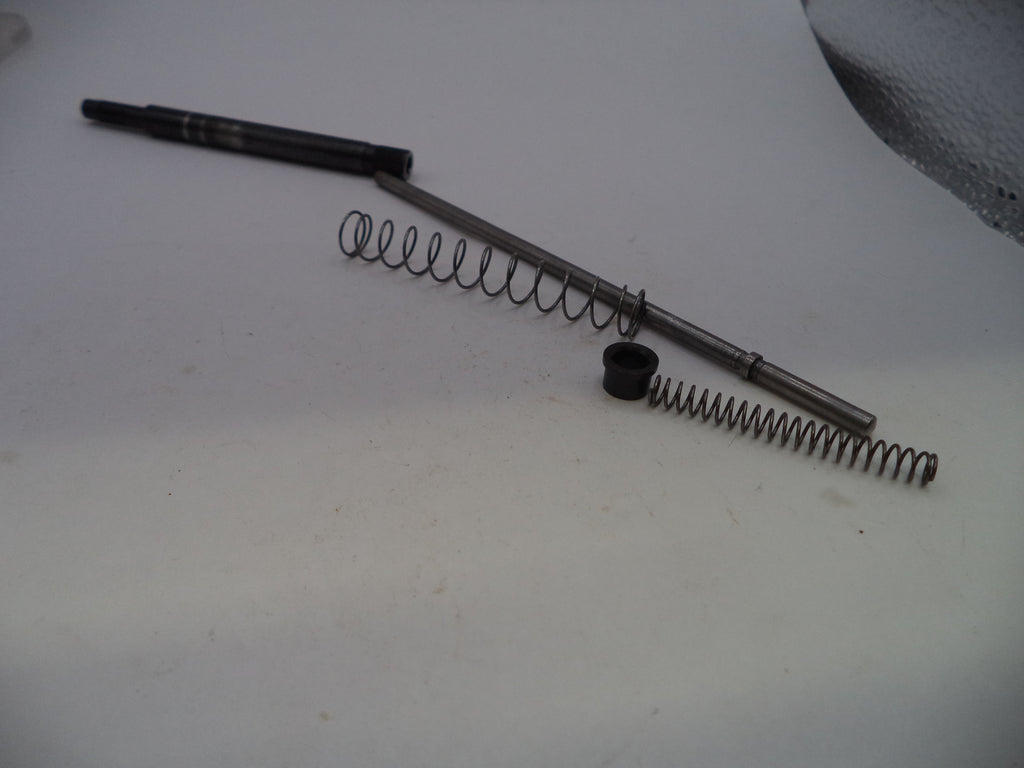A set of mechanical parts including a long metal rod, two coiled metal springs, and a small black plastic cap, arranged on a plain off-white surface.