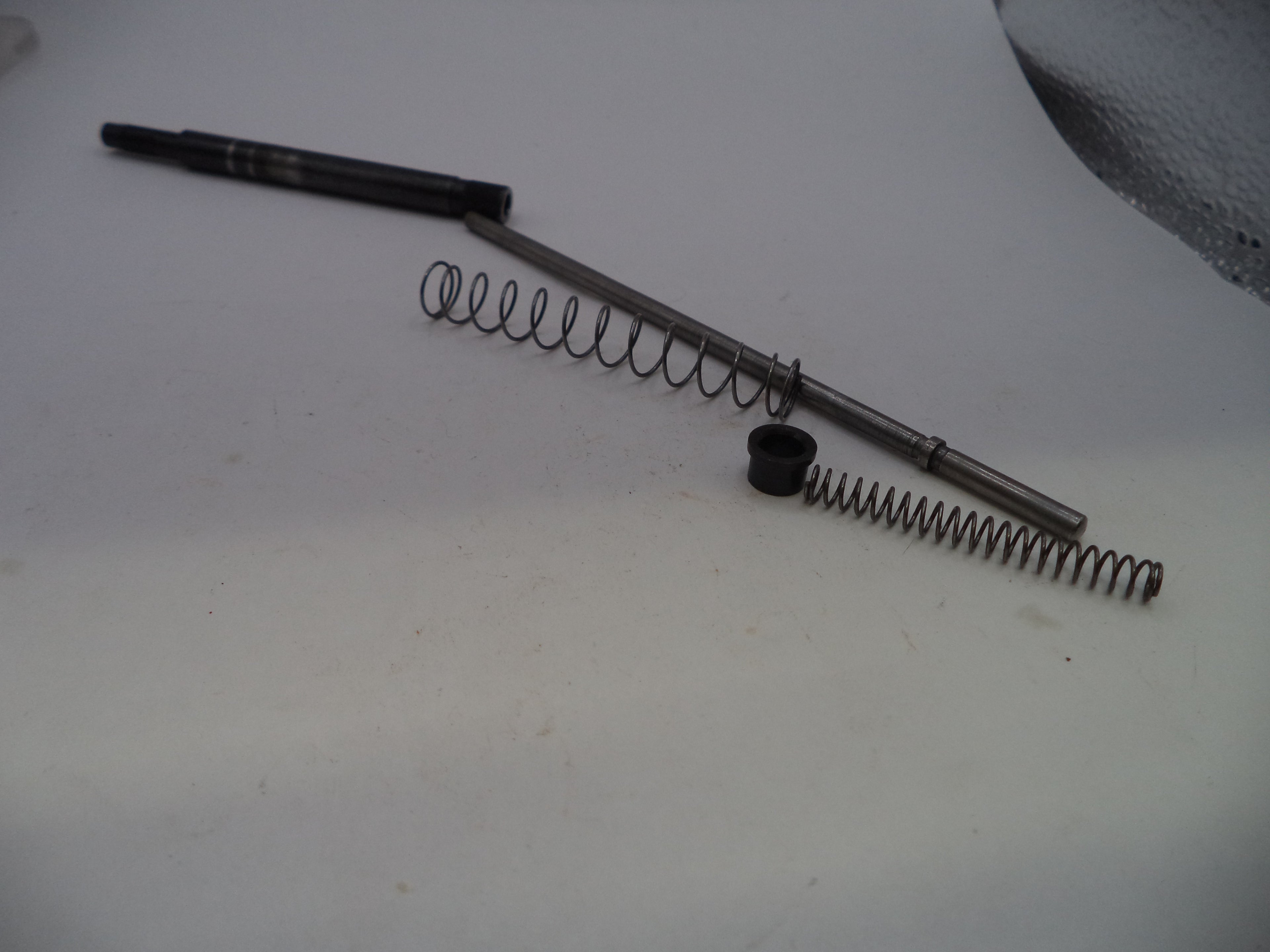 A set of mechanical parts including a long metal rod, two coiled metal springs, and a small black plastic cap, arranged on a plain off-white surface.