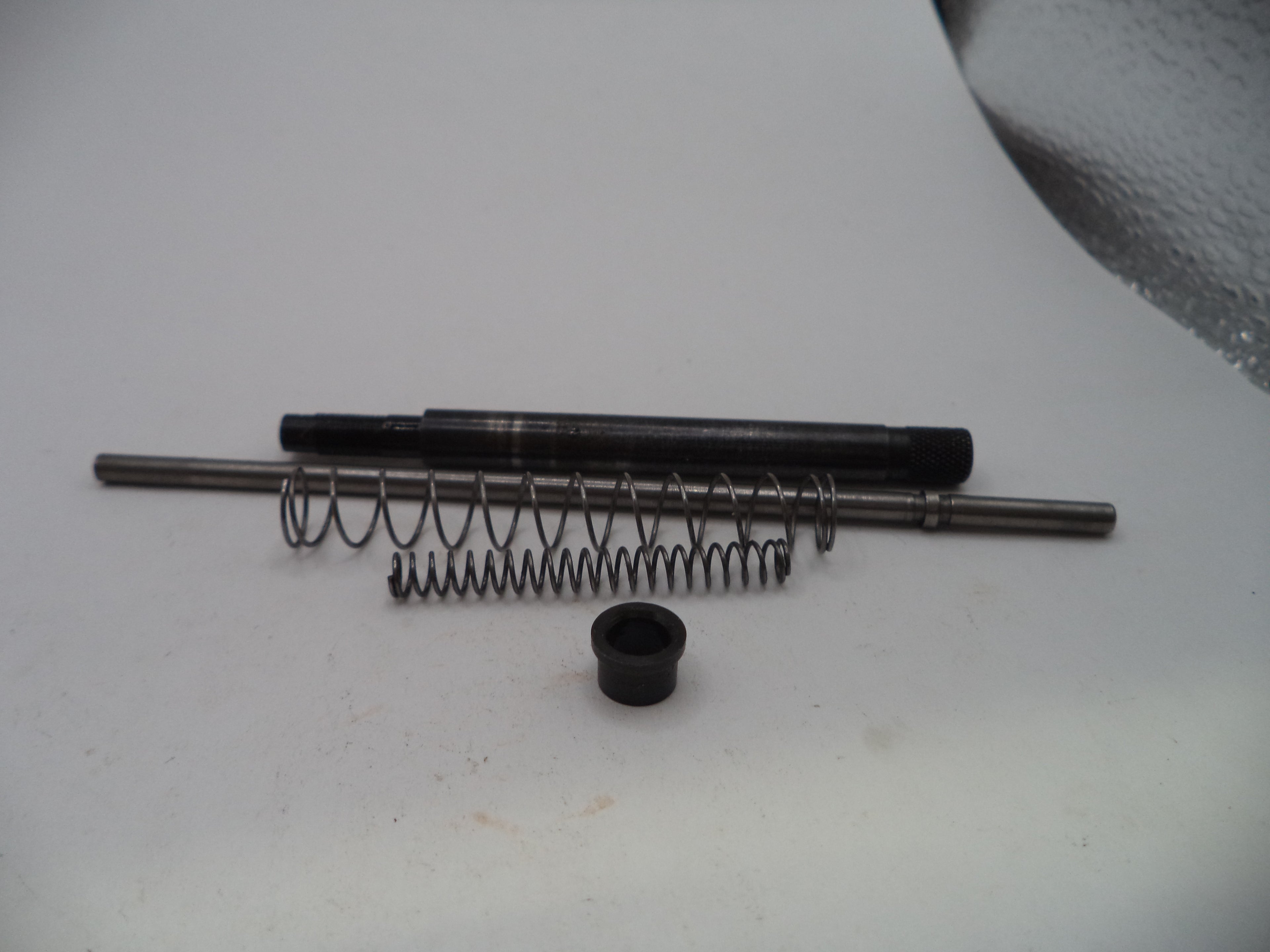 A set of small mechanical parts including a black metal rod, a coiled metal spring, a small black cylindrical piece, and a long silver metal rod, arranged on a white surface.