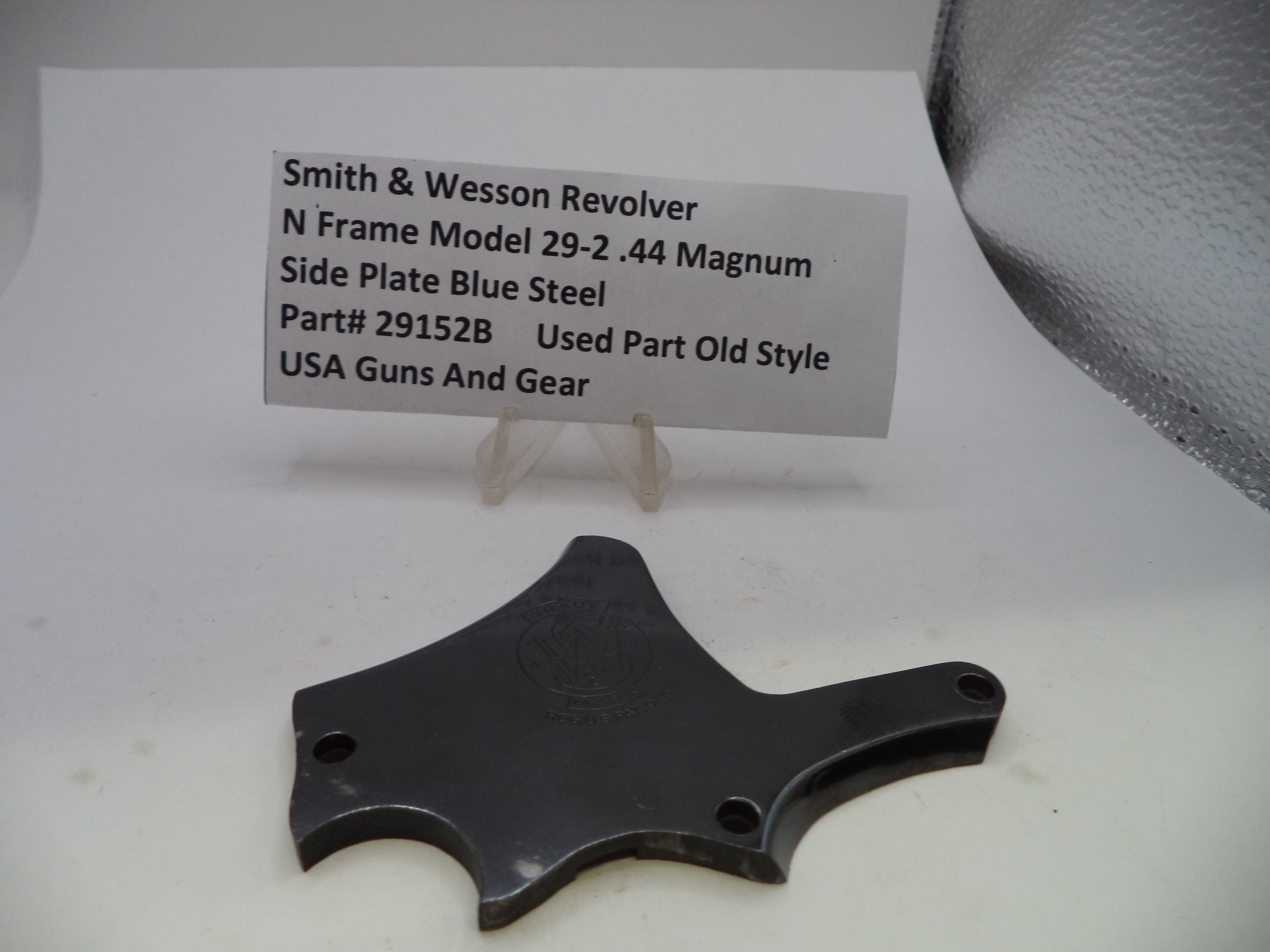 A used black metal side plate for a Smith & Wesson revolver, displayed on a white surface with a small label card showing product details.