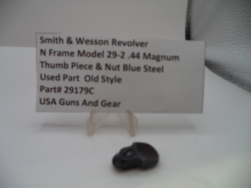 A small black metal thumb piece for a Smith & Wesson revolver, displayed on a clear plastic stand next to a white label card with product details.