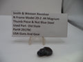 A small black metal thumb piece for a Smith & Wesson revolver, displayed on a clear plastic stand next to a white label card with product details.