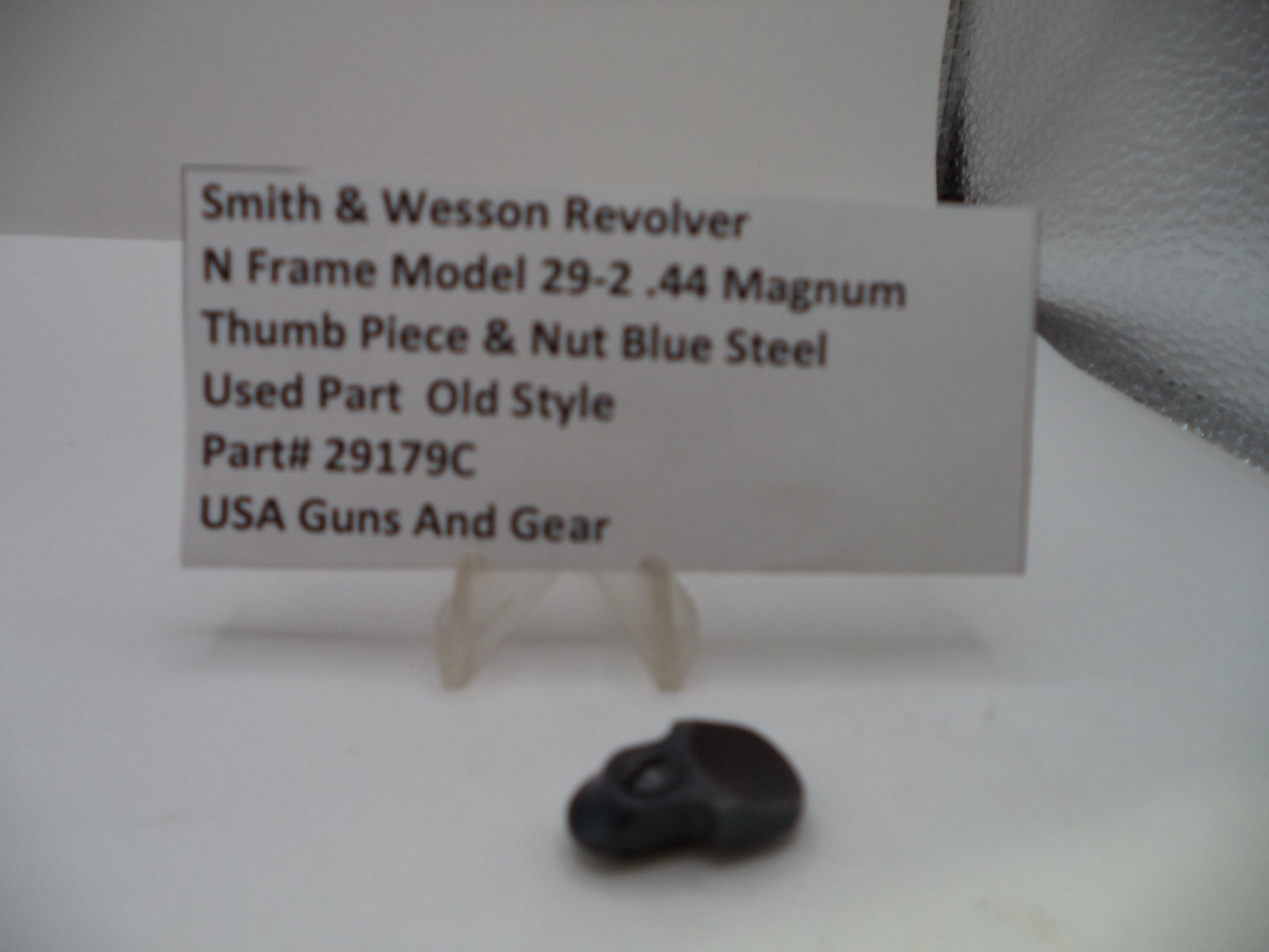 A small black metal thumb piece for a Smith & Wesson revolver, displayed on a clear plastic stand next to a white label card with product details.