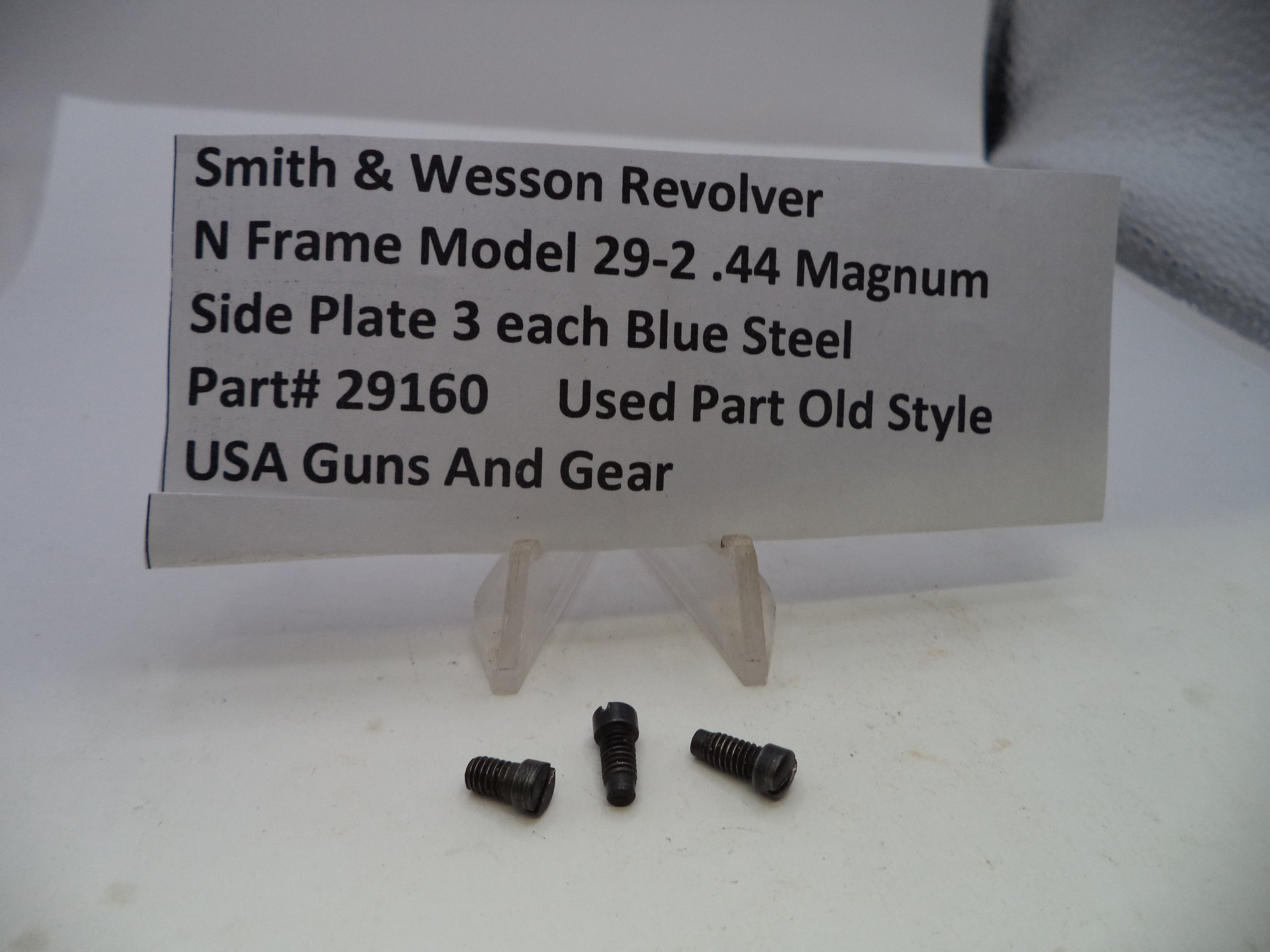 Three small dark metal screws displayed in front of a white sign that reads 'Smith & Wesson Revolver N Frame Model 29-2 .44 Magnum Side Plate 3 each Blue Steel Part# 29160 Used Part Old Style USA Guns And Gear' on a white surface.