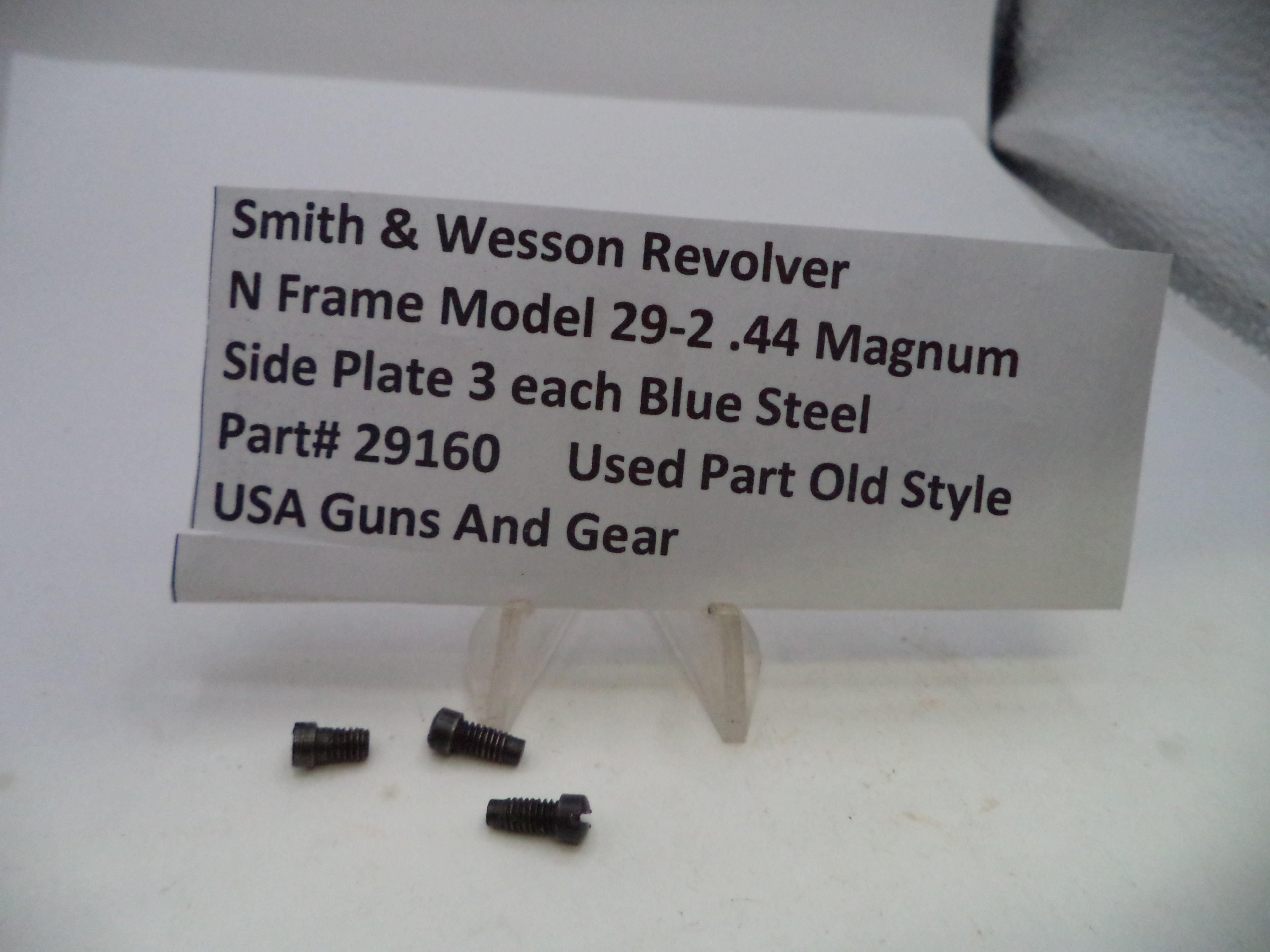 Three small dark metal screws displayed in front of a white sign that reads 'Smith & Wesson Revolver N Frame Model 29-2 .44 Magnum Side Plate 3 each Blue Steel Part# 29160 Used Part Old Style USA Guns And Gear' on a white surface.