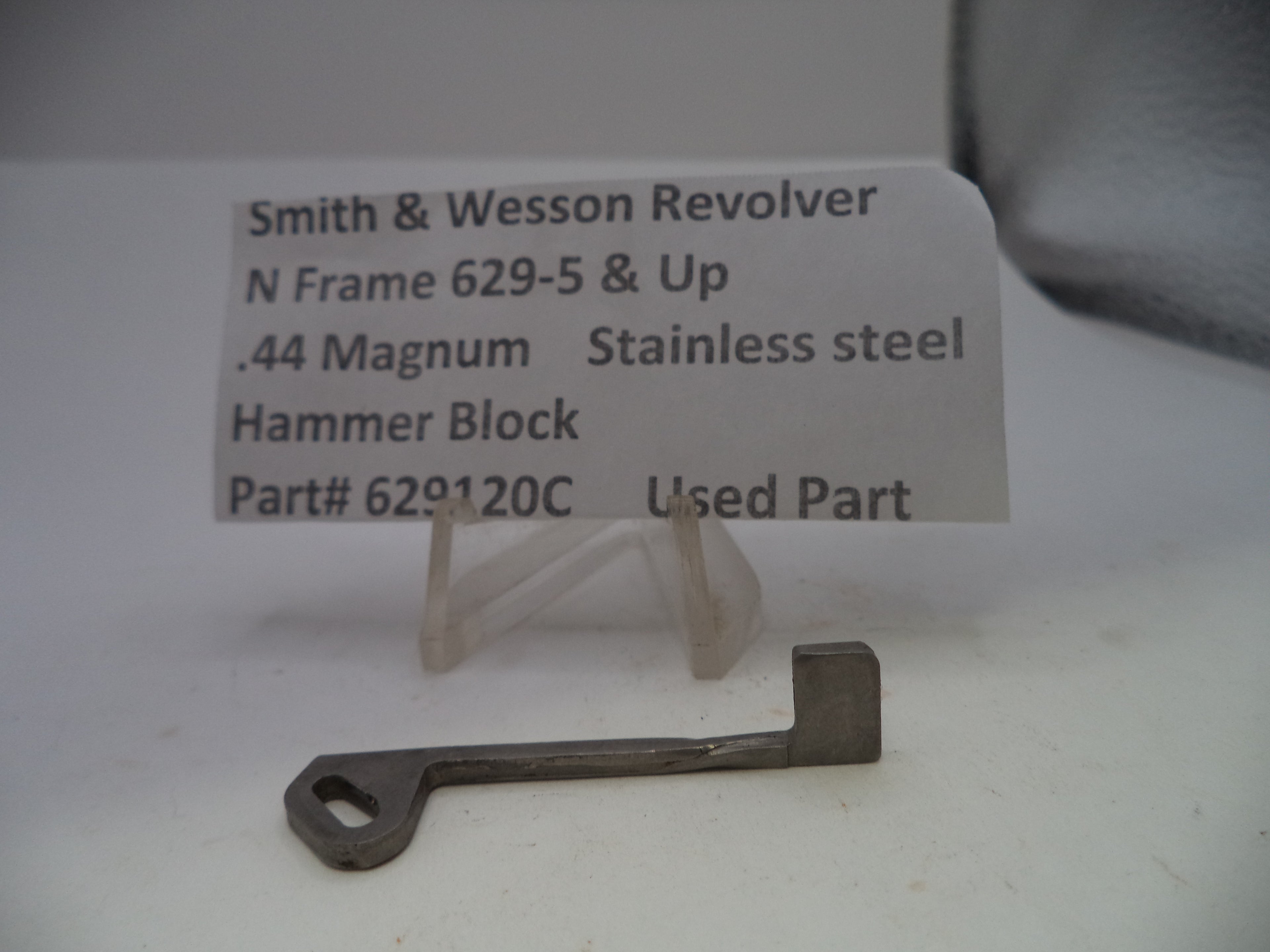 A small metal used part for a Smith & Wesson revolver, displayed on a clear plastic stand in front of a white label card with product details.