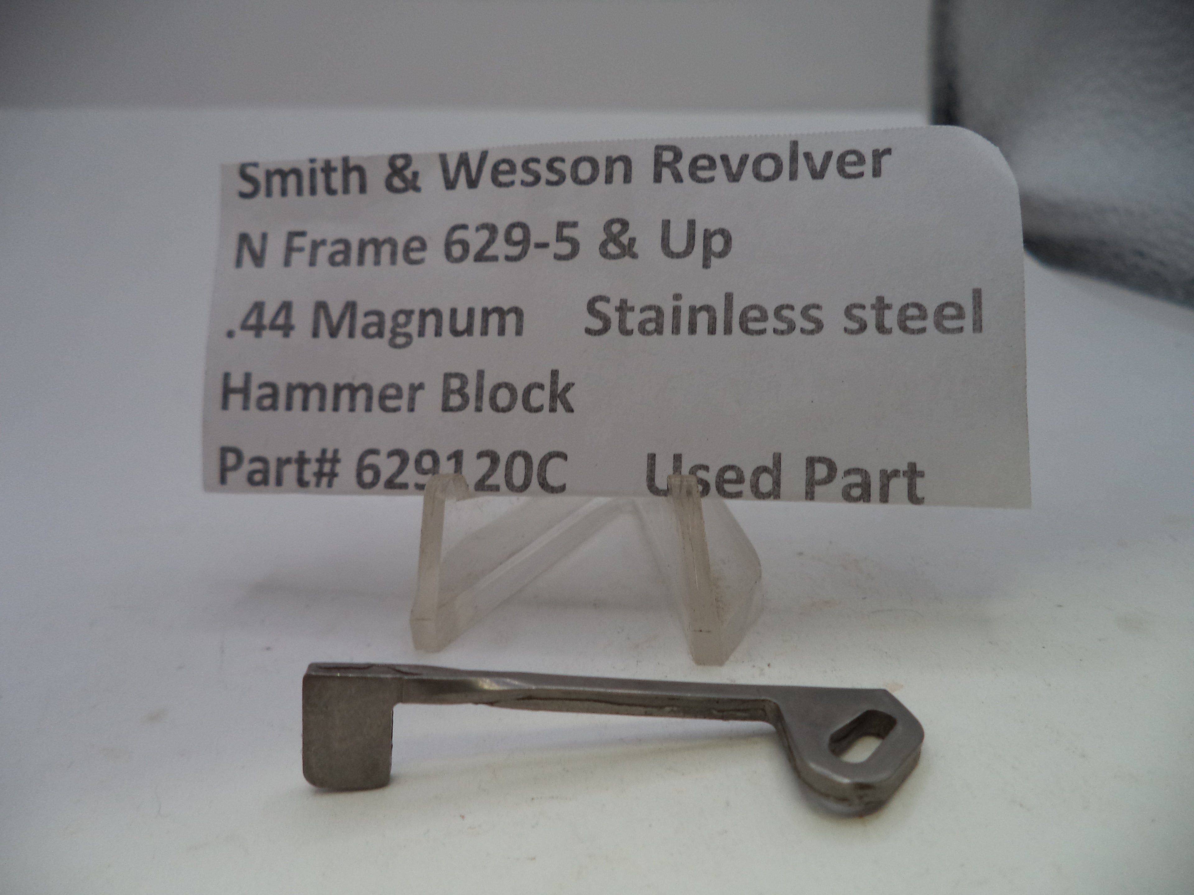 A small metal hammer block part displayed on a clear plastic stand in front of a printed label that reads 'Smith & Wesson Revolver N Frame 629-5 & Up .44 Magnum Stainless steel Hammer Block Part# 629120C Used Part'.