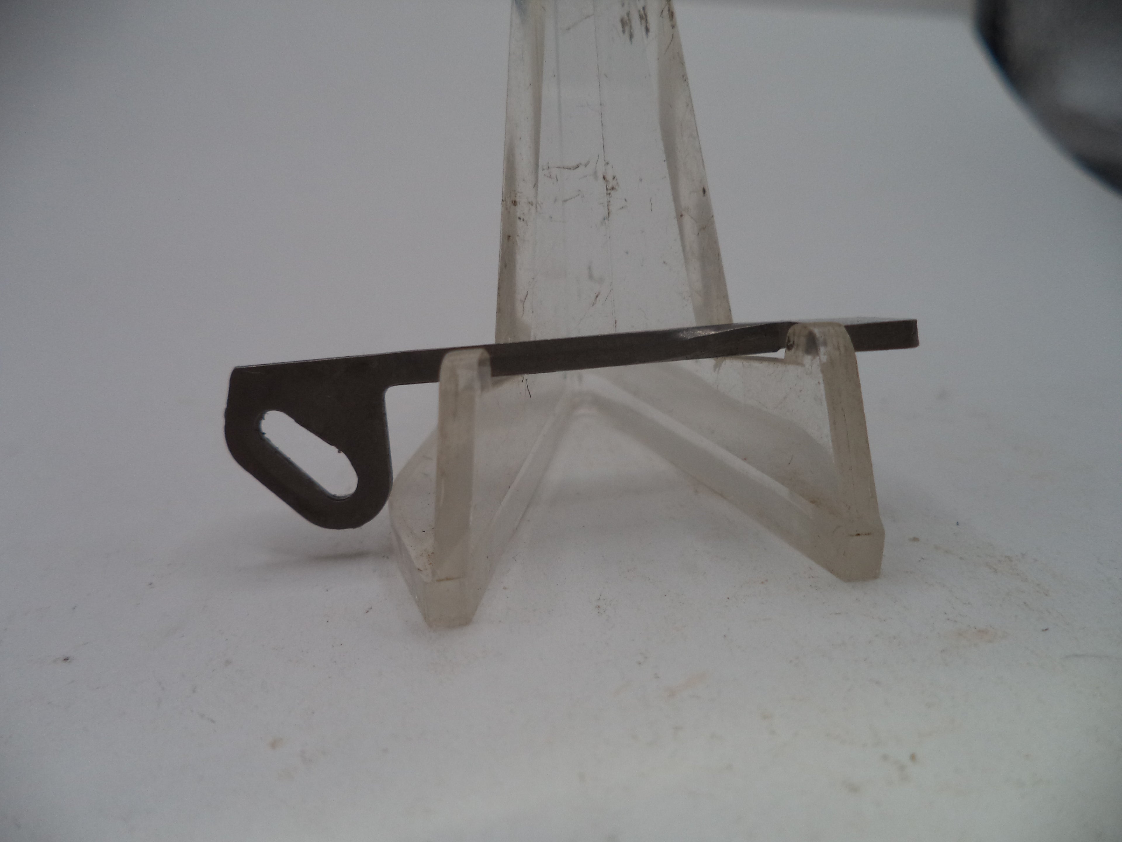 A small flat metal tool or blade with a looped end, displayed on a clear plastic stand against a plain light background.