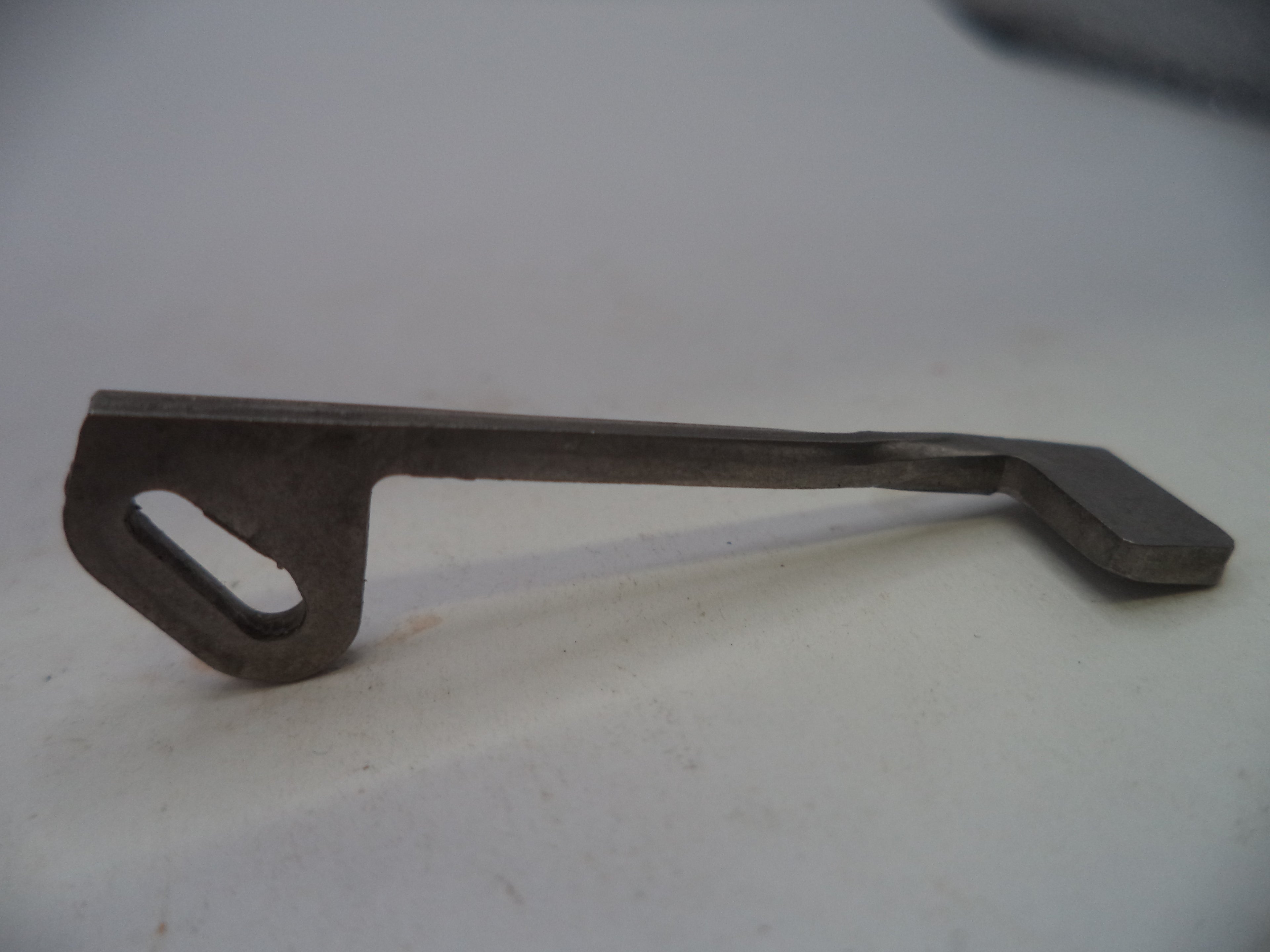 A small dark metal tool or hardware piece with a flat rectangular head and a looped handle, photographed on a plain light background.