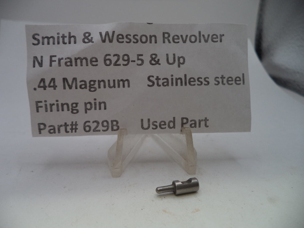 A small metal firearm firing pin part displayed on a clear plastic stand in front of a white paper label that reads 'Smith & Wesson Revolver N Frame 629-5 & Up .44 Magnum Stainless steel Firing pin Part# 629B Used Part'.