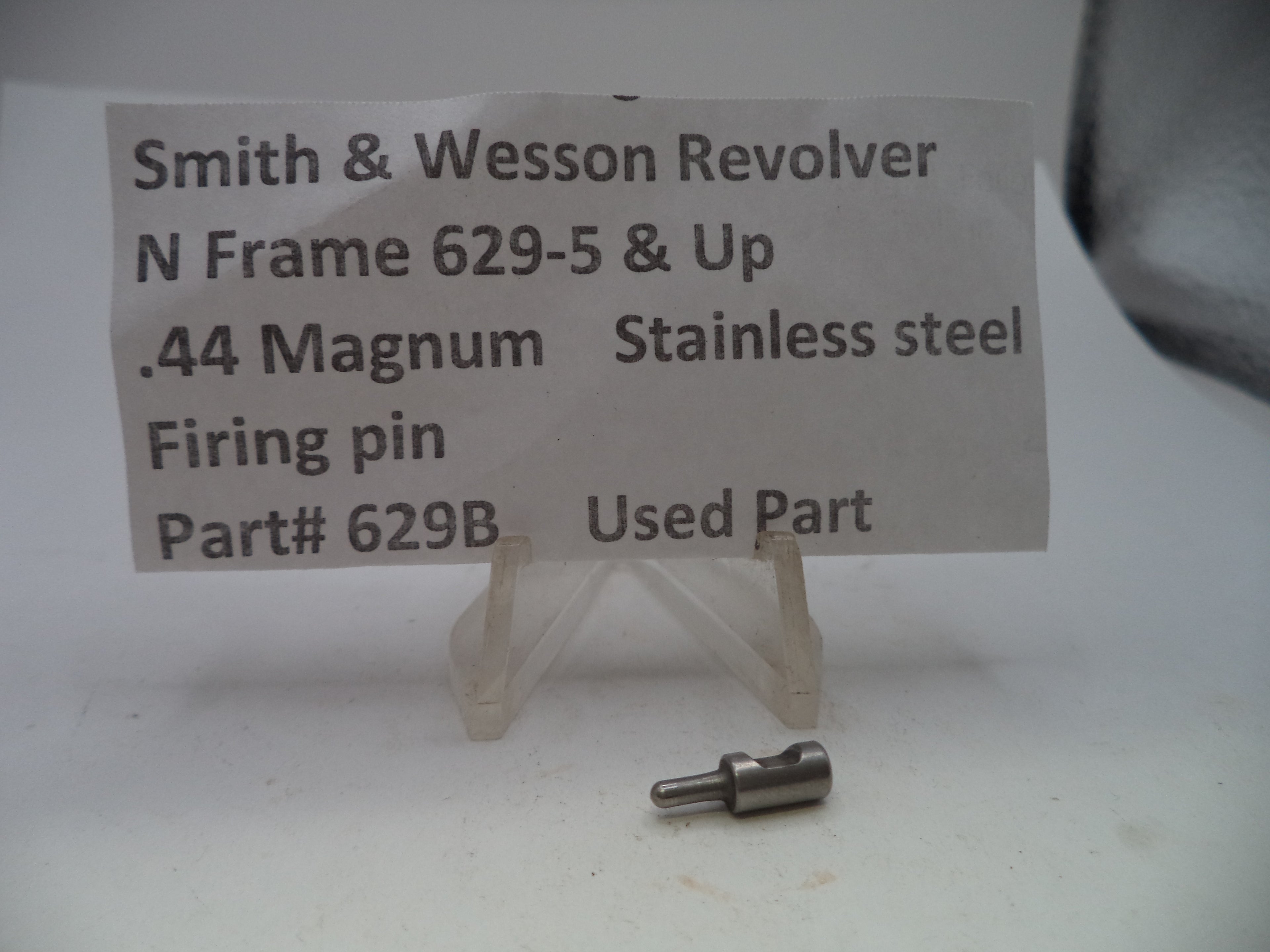 A small metal firearm firing pin part displayed on a clear plastic stand in front of a white paper label that reads 'Smith & Wesson Revolver N Frame 629-5 & Up .44 Magnum Stainless steel Firing pin Part# 629B Used Part'.