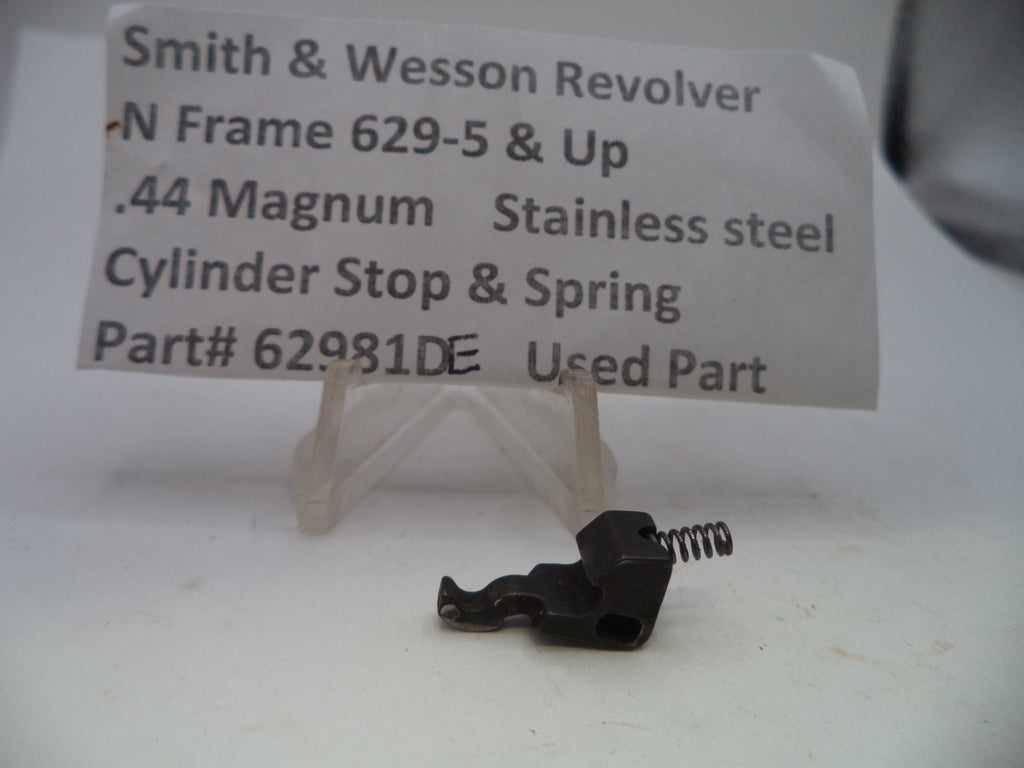 A small black metal cylinder stop and spring part for a Smith & Wesson revolver, displayed on a clear plastic stand with a paper label in the background.