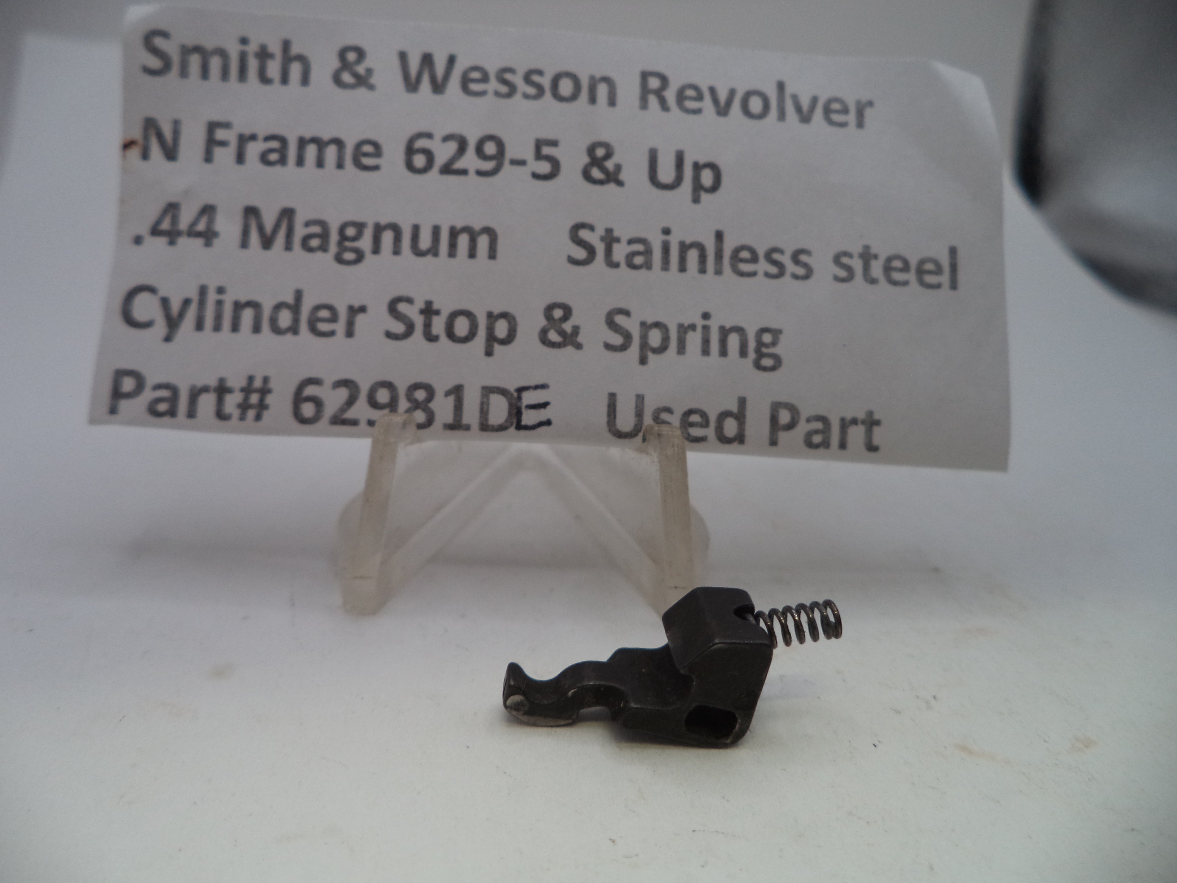 A small black metal cylinder stop and spring part for a Smith & Wesson revolver, displayed on a clear plastic stand with a paper label in the background.