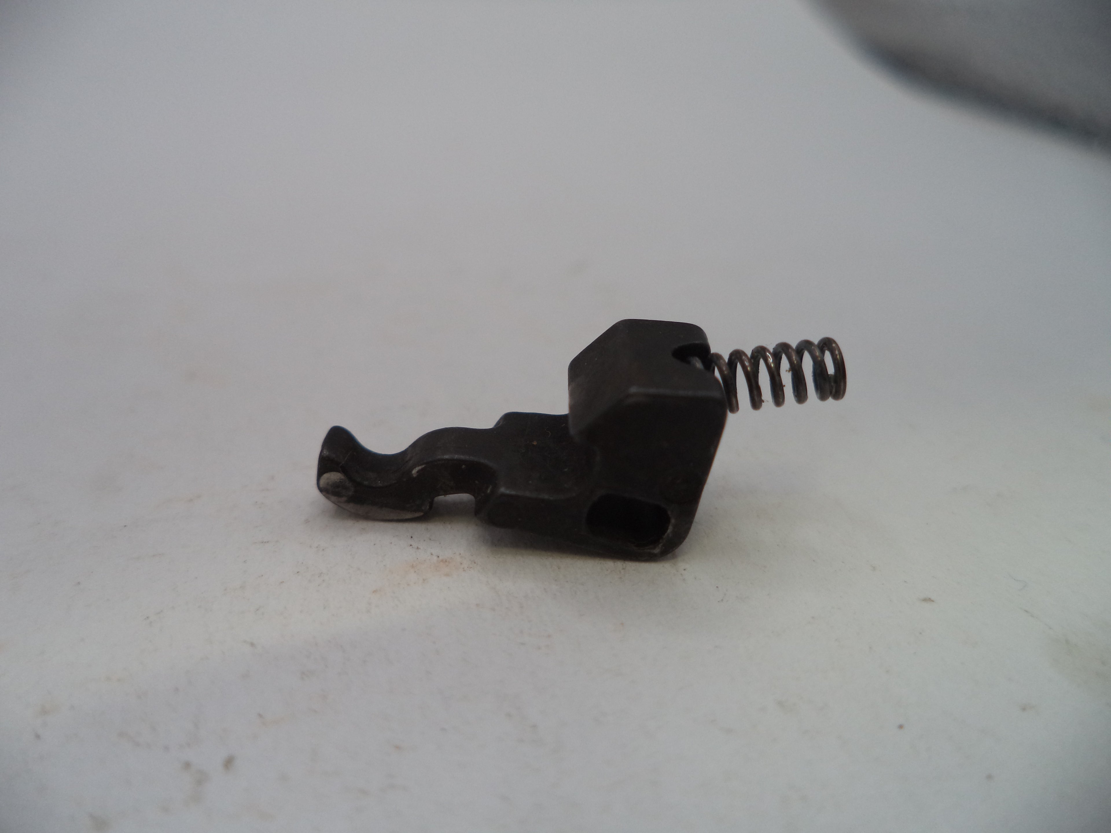 A small black metal mechanical component with a coiled spring, photographed on a plain off-white surface.