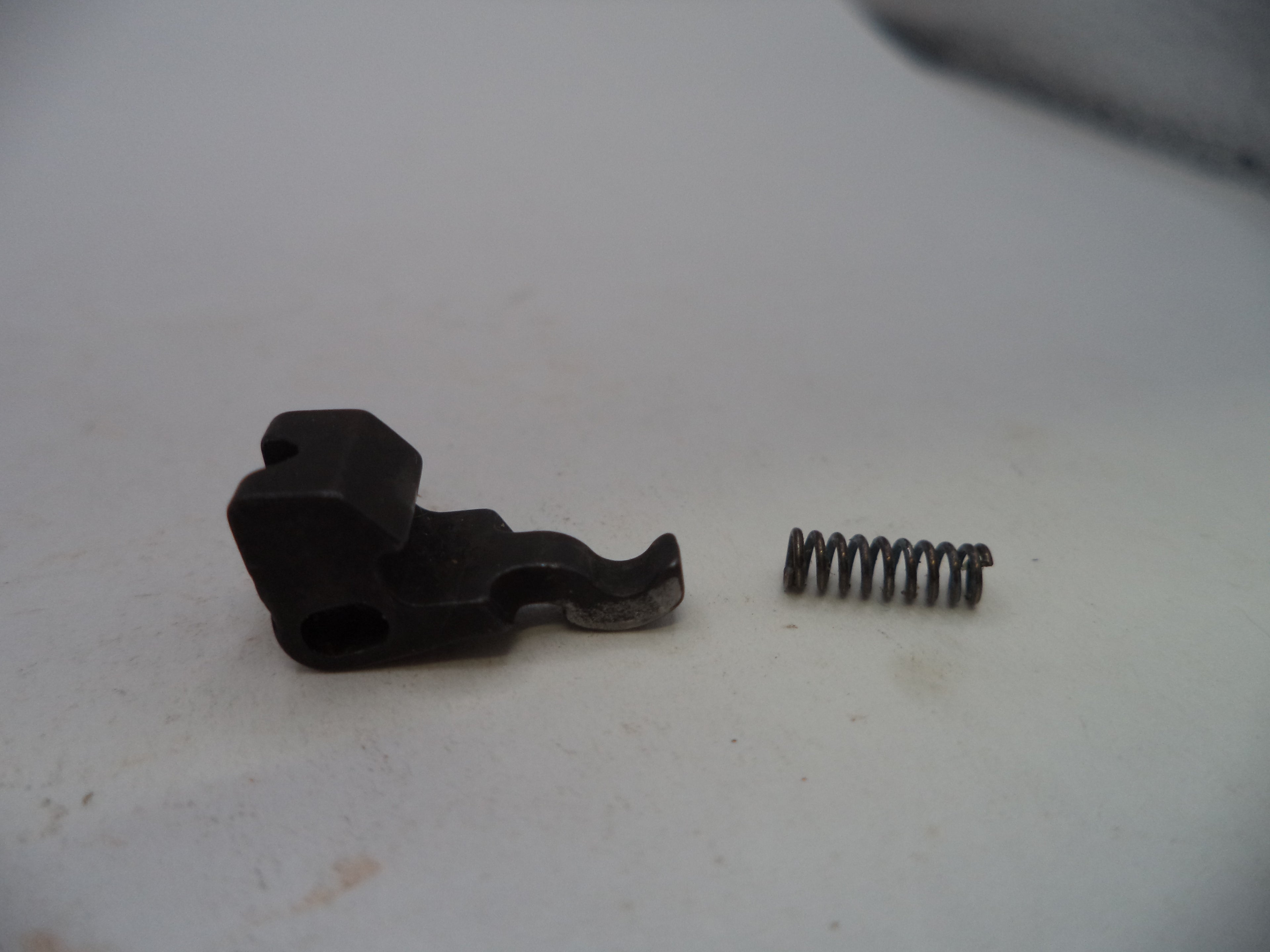 A small black metal mechanical component with a coiled spring next to it, photographed on a plain off-white surface.