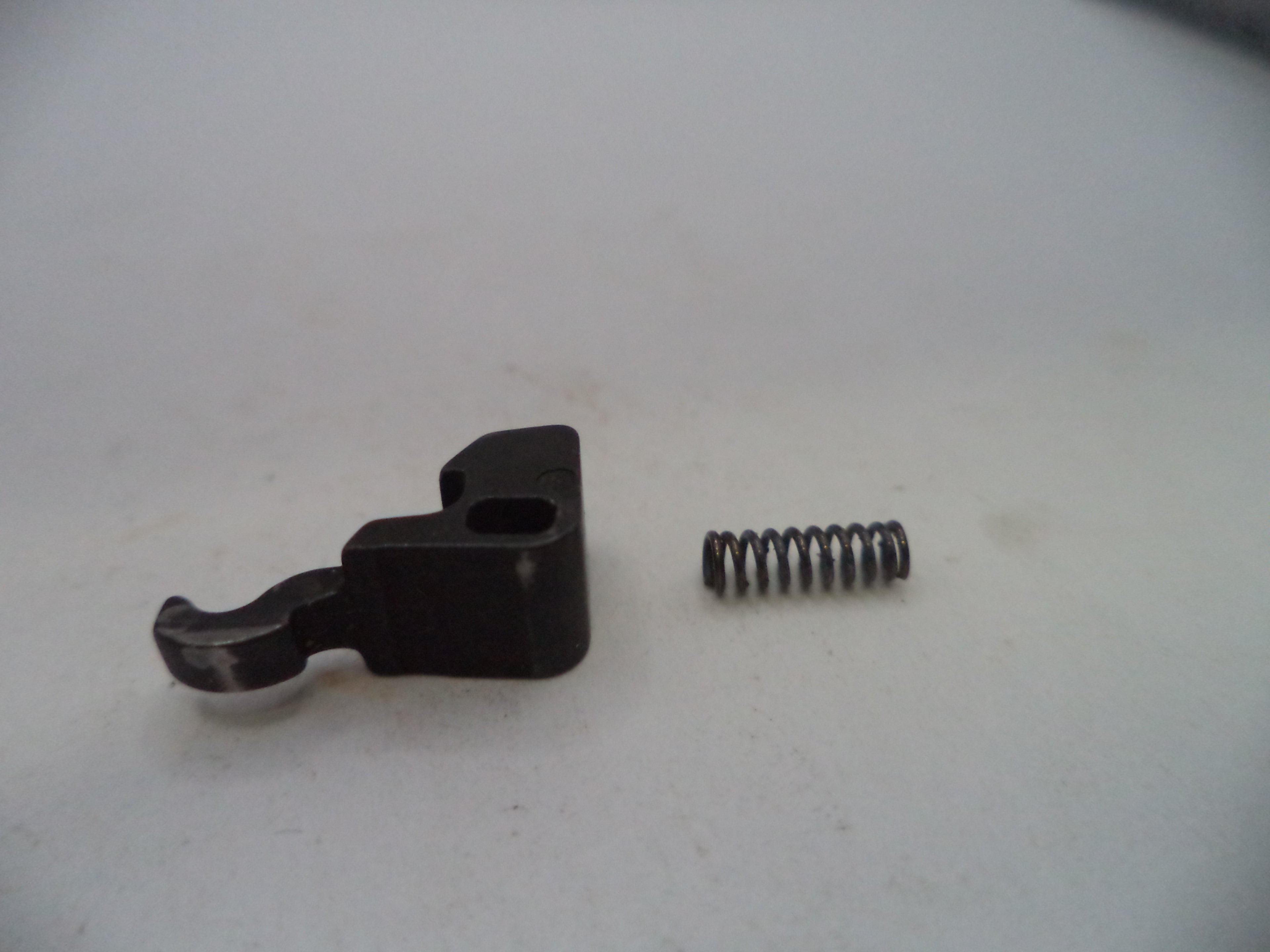 A small black metal mechanical component with a coiled metal spring next to it, photographed on a plain light gray surface.