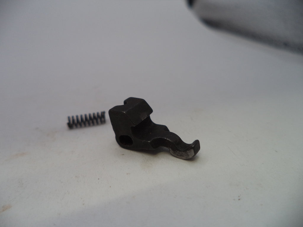 A small black metal mechanical component with a small silver coil spring beside it, photographed on a plain off-white surface.