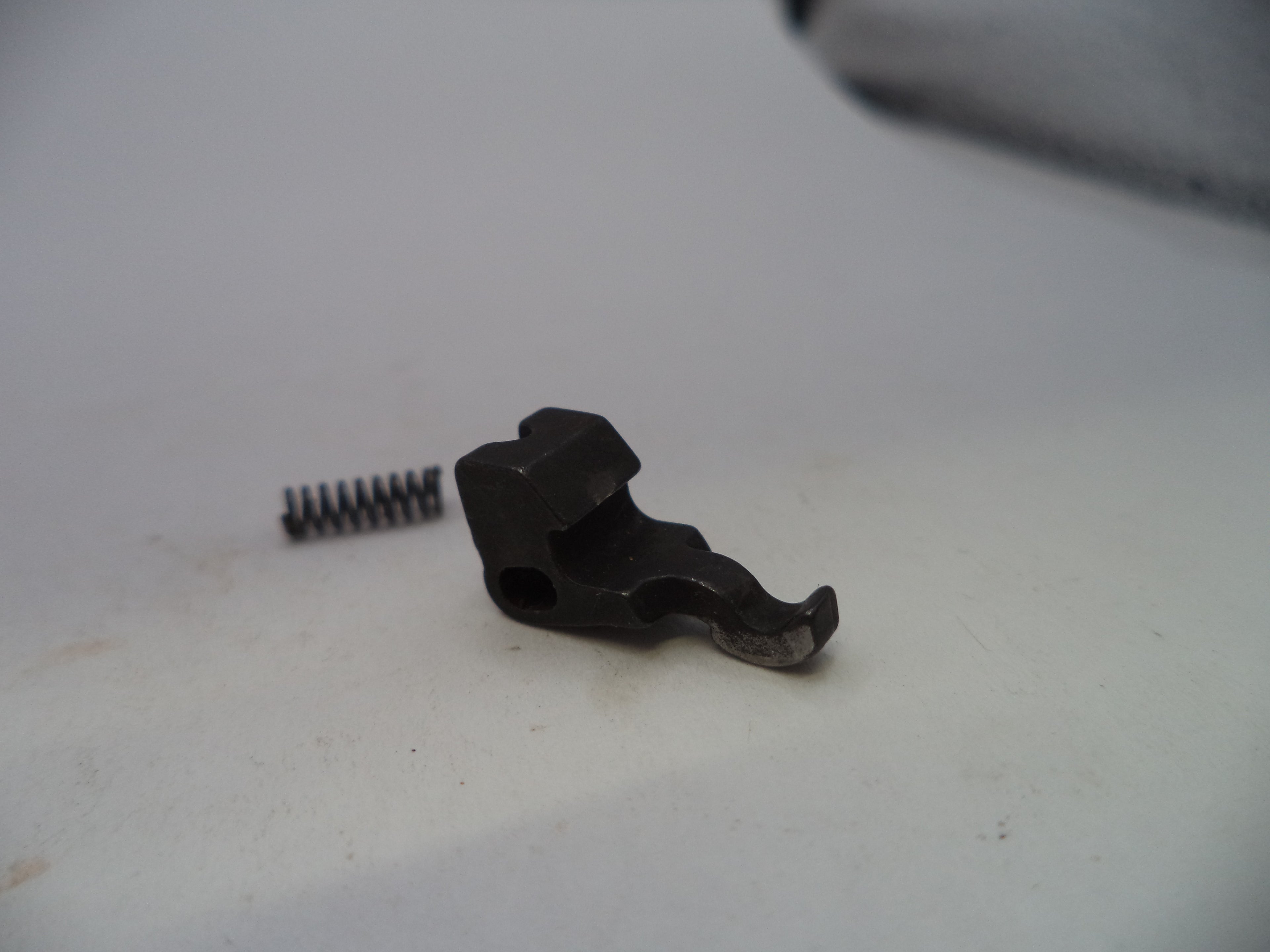 A small black metal mechanical component with a small silver coil spring beside it, photographed on a plain off-white surface.