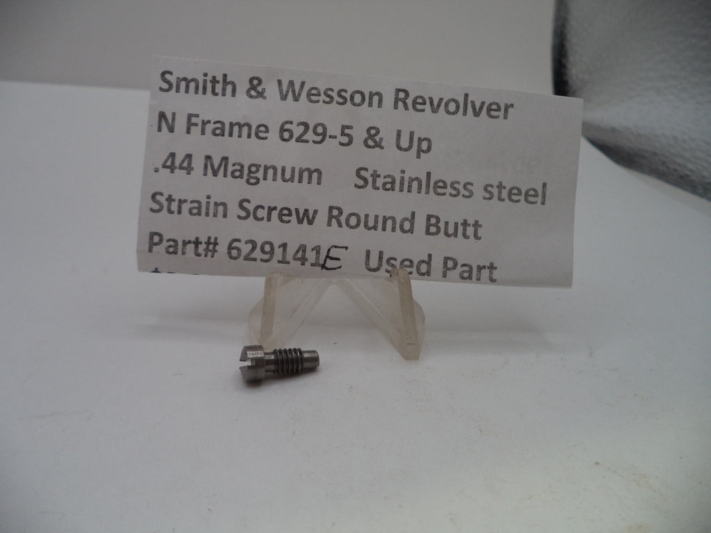 A small metal strain screw part displayed on a clear plastic stand in front of a white paper label that reads 'Smith & Wesson Revolver N Frame 629-5 & Up .44 Magnum Stainless steel Strain Screw Round Butt Part# 629141E Used Part'.