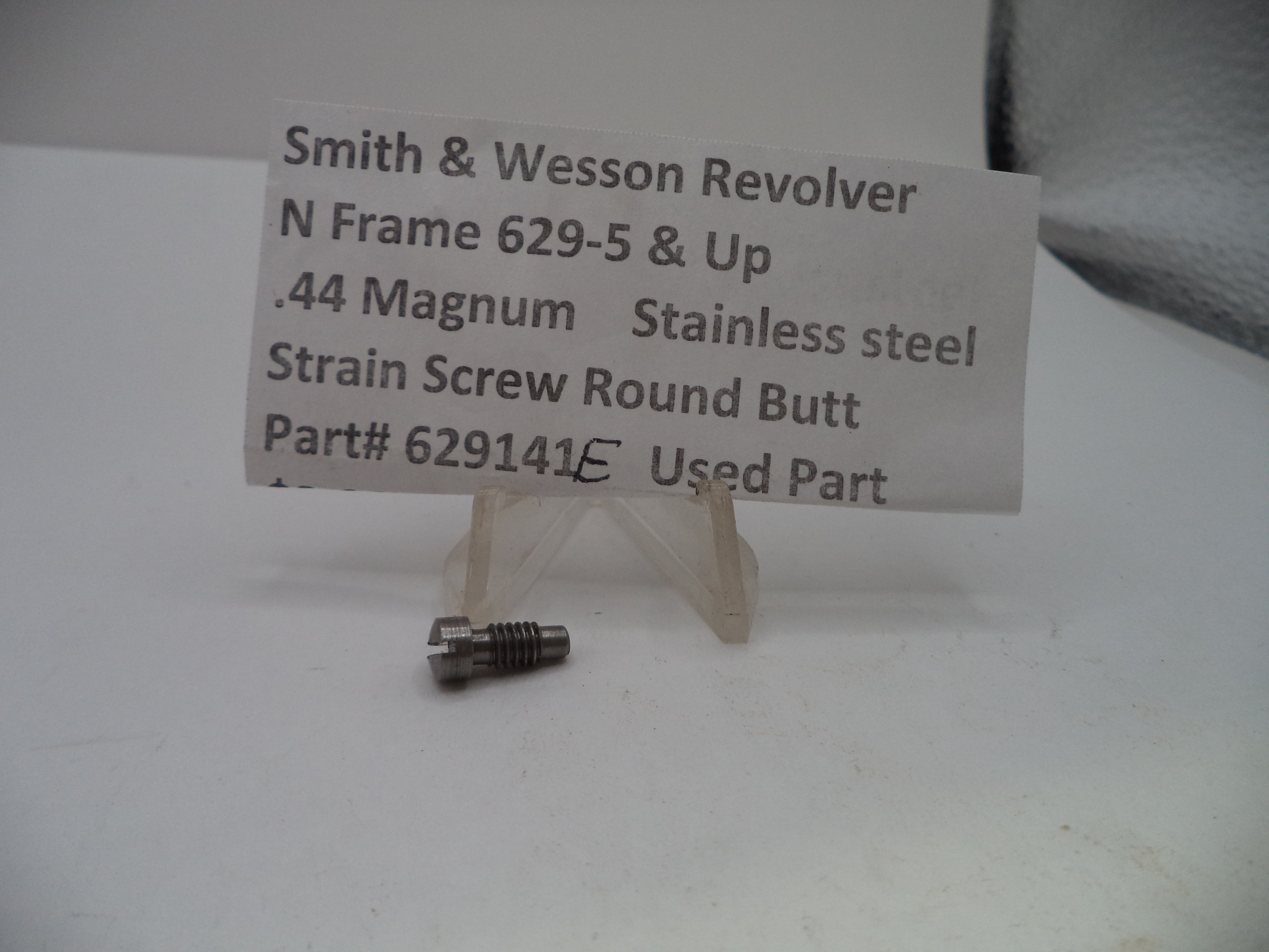 A small metal strain screw part displayed on a clear plastic stand in front of a white paper label that reads 'Smith & Wesson Revolver N Frame 629-5 & Up .44 Magnum Stainless steel Strain Screw Round Butt Part# 629141E Used Part'.