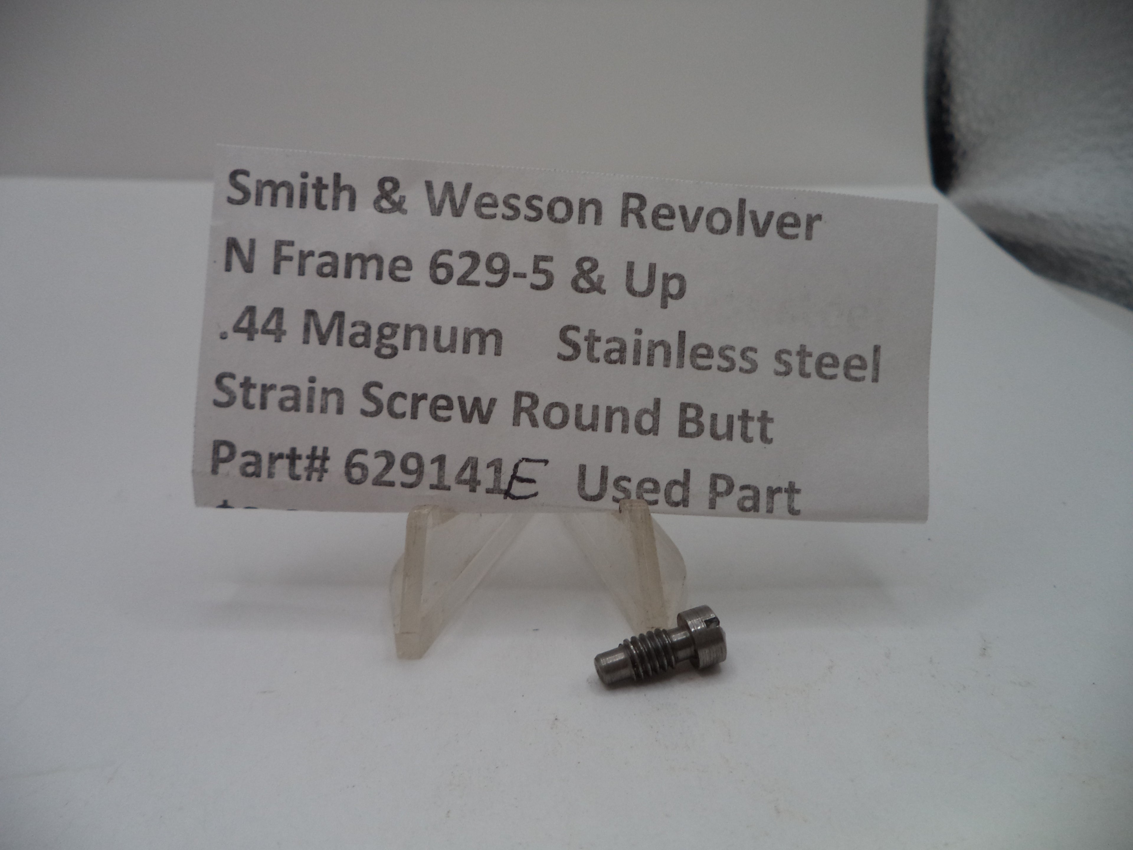 A small metal strain screw part displayed on a clear plastic stand with a handwritten label card that reads 'Smith & Wesson Revolver N Frame 629-5 & Up .44 Magnum Stainless steel Strain Screw Round Butt Part# 629141 Used Part'.