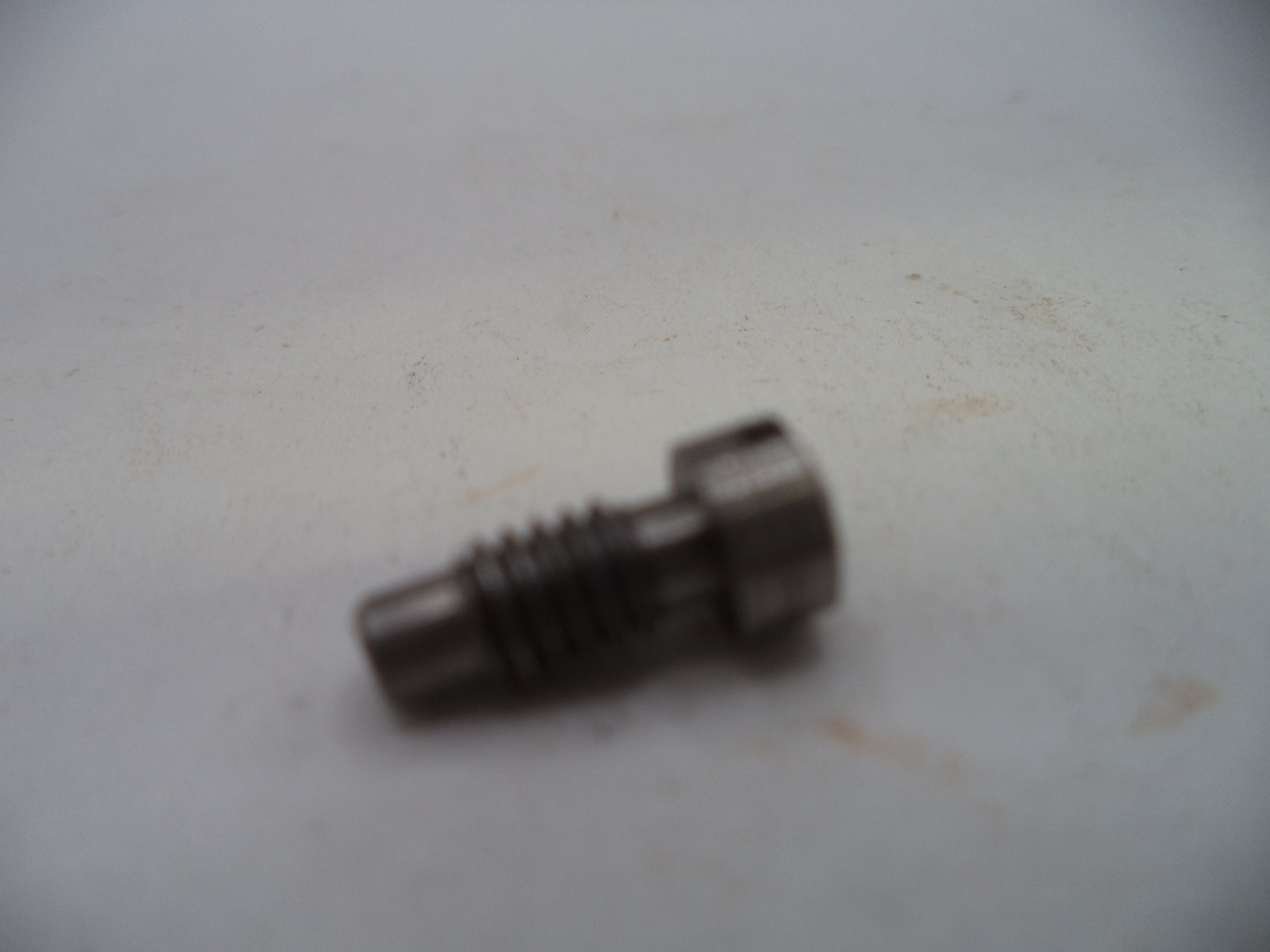 A small metallic threaded component, possibly a bolt or pin, photographed on a plain off-white surface.