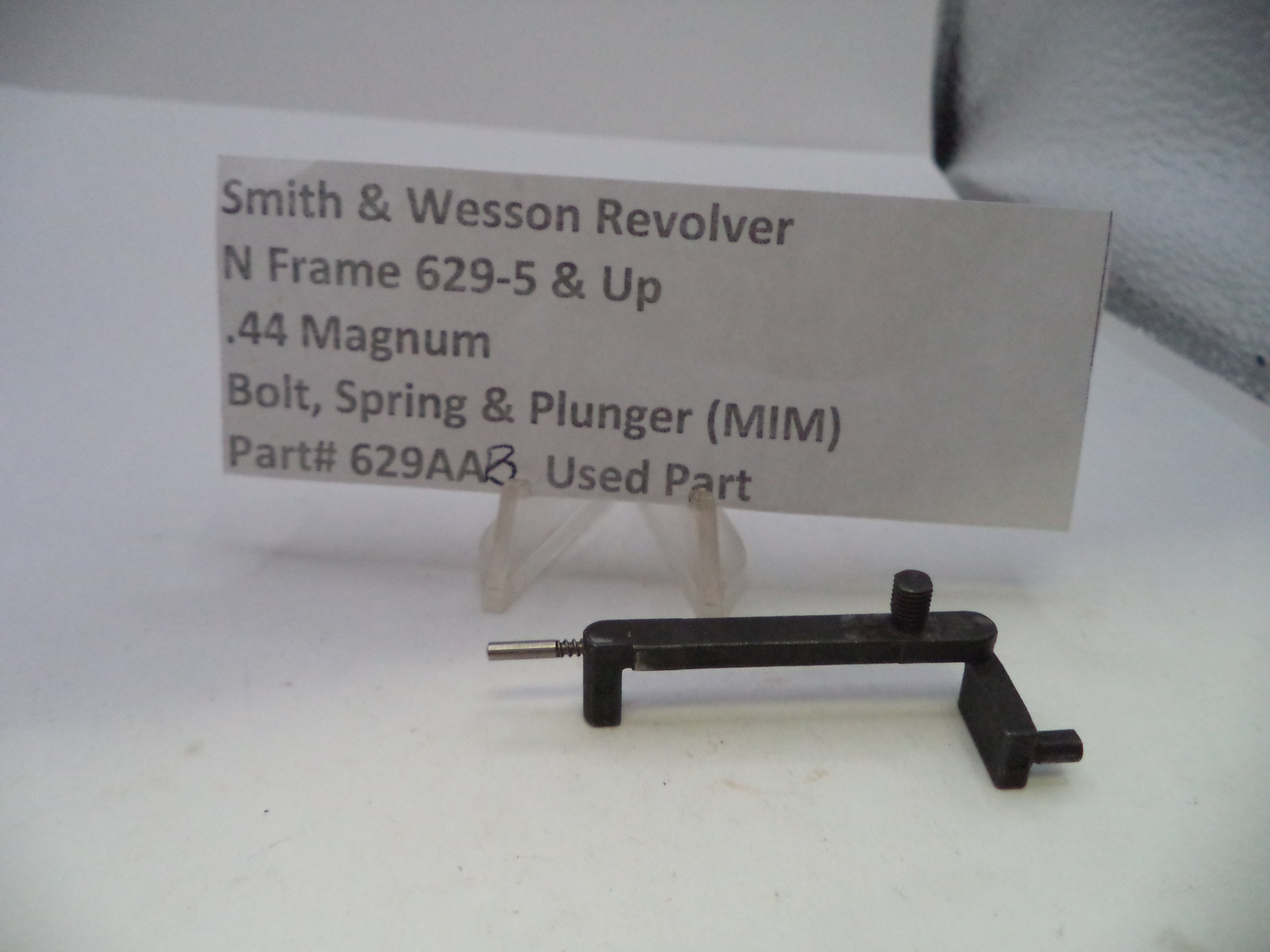 A used black metal Smith & Wesson revolver bolt, spring, and plunger part, displayed on a small clear stand in front of a paper label card.