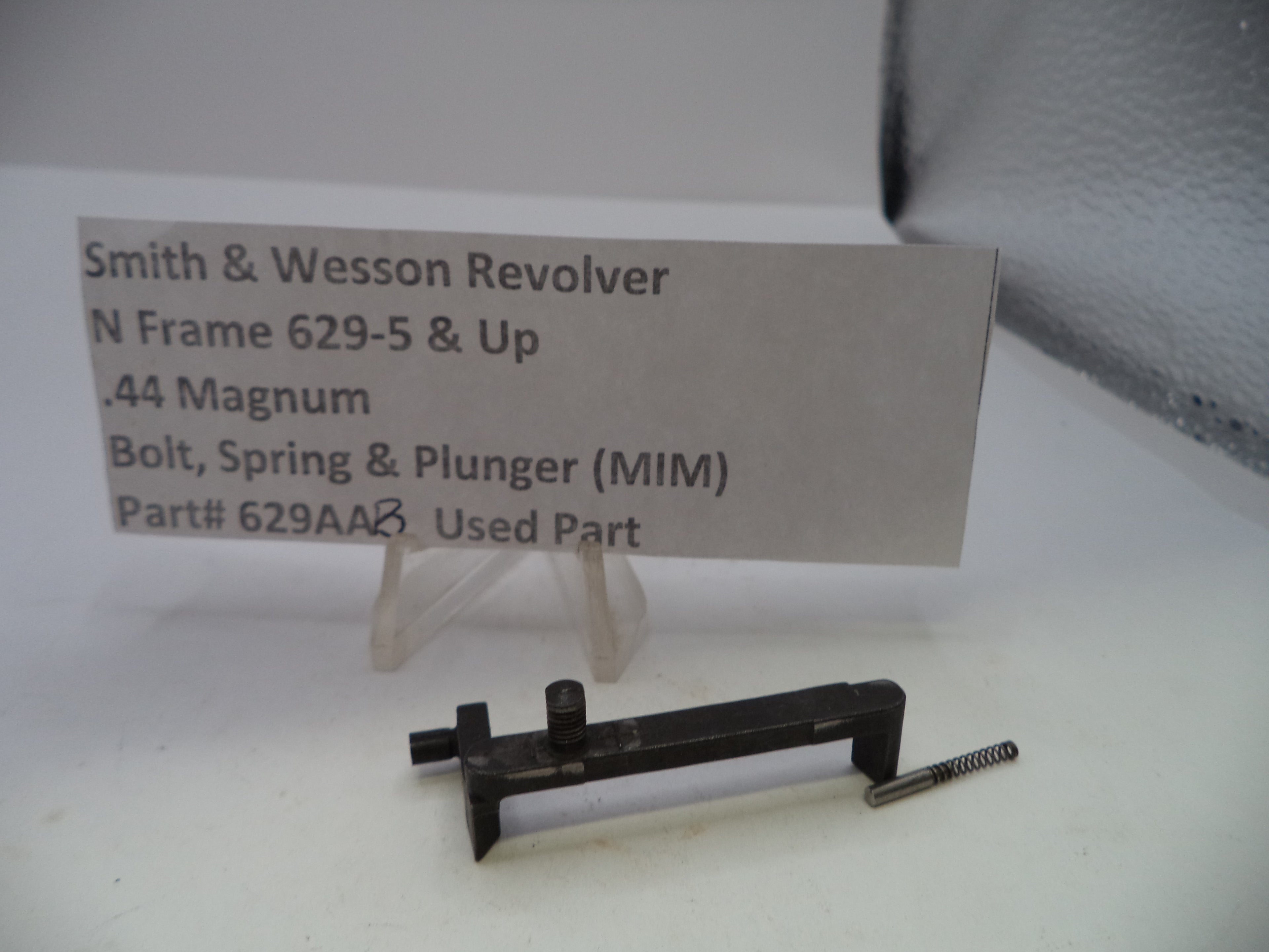 A used Smith & Wesson revolver bolt, spring, and plunger part shown on a white surface with a product information card behind it. The part is black metal with a small silver spring.