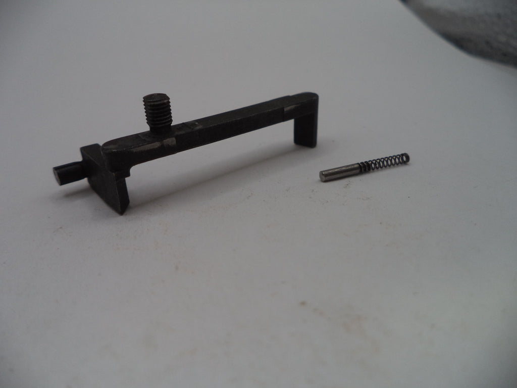 A black metal trigger assembly component with a threaded screw post and a small separate metal spring, photographed on a plain light gray surface.