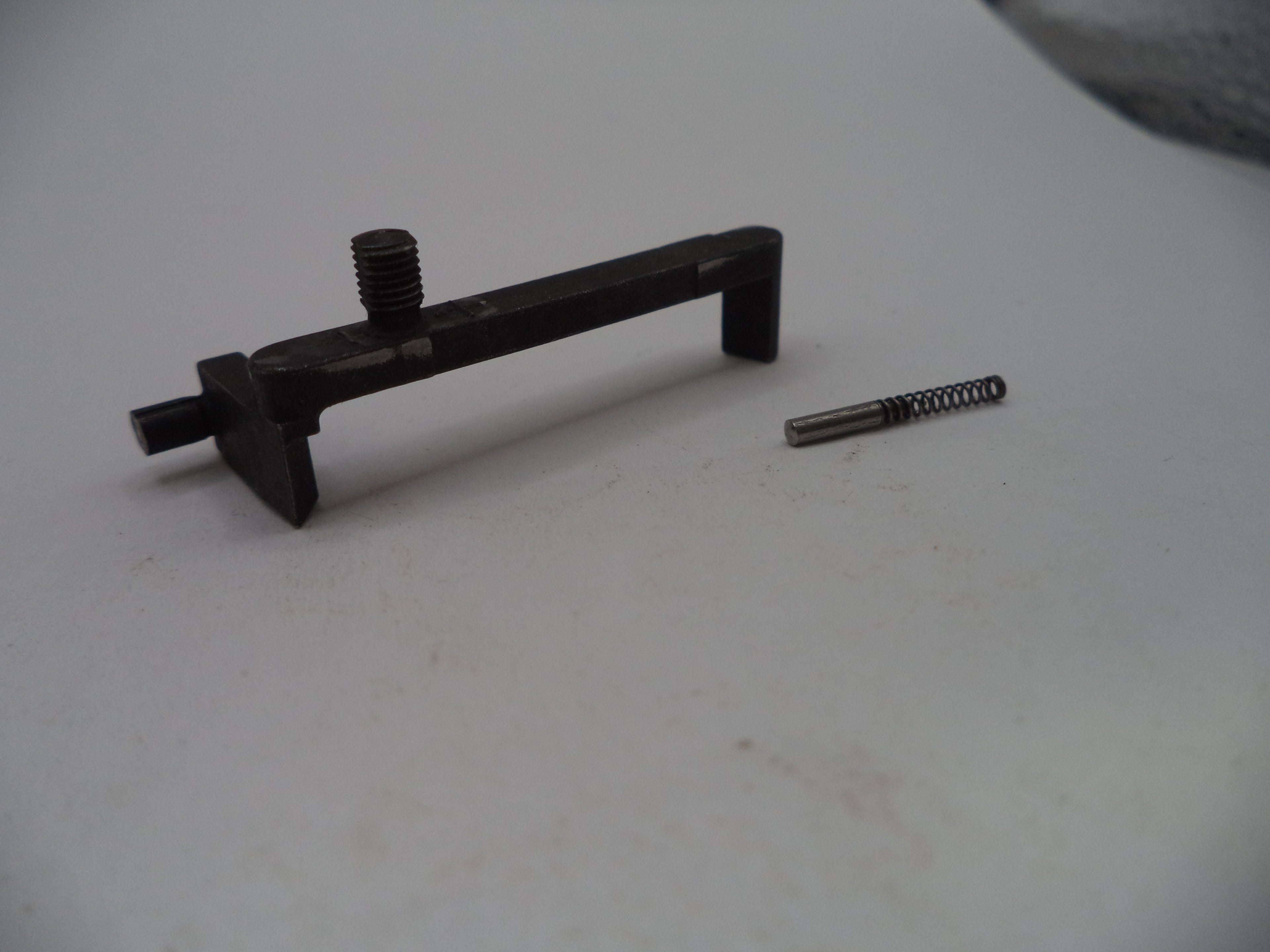 A black metal trigger assembly component with a threaded screw post and a small separate metal spring, photographed on a plain light gray surface.