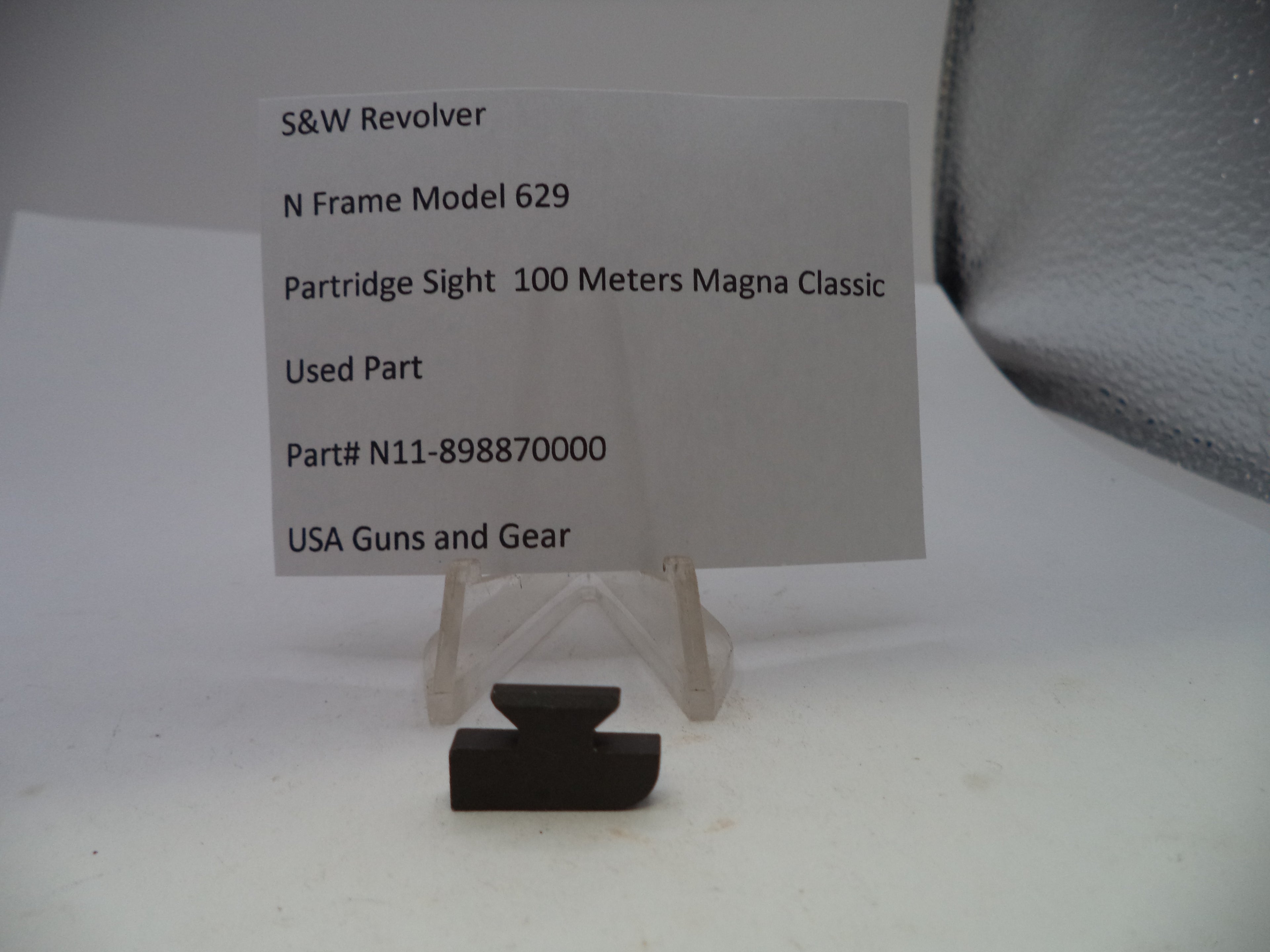 A small black used part for an S&W Revolver N Frame Model 629 partridge sight, displayed on a clear plastic stand next to a paper label card with part details.