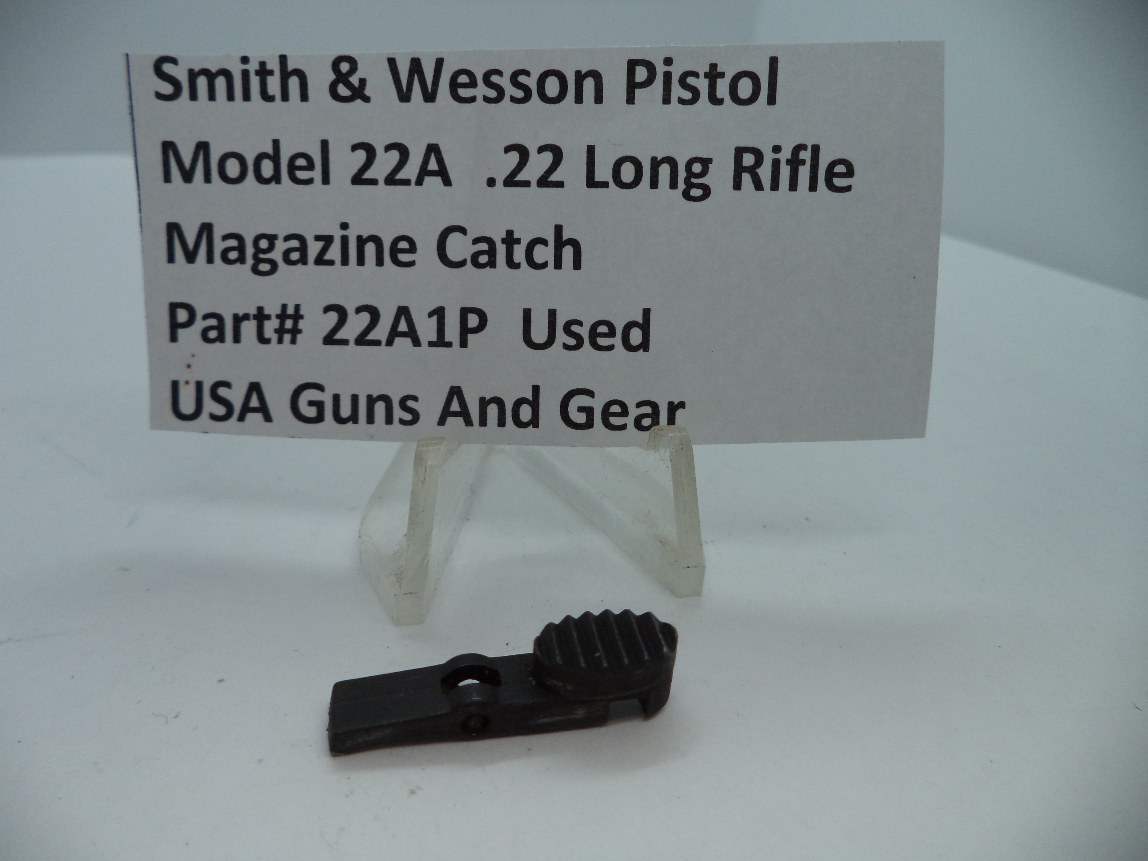 Smith & Wesson Pistol Model 22A Magazine Catch .22 Long Rifle Used Part displayed on a white stand with a label providing product details.