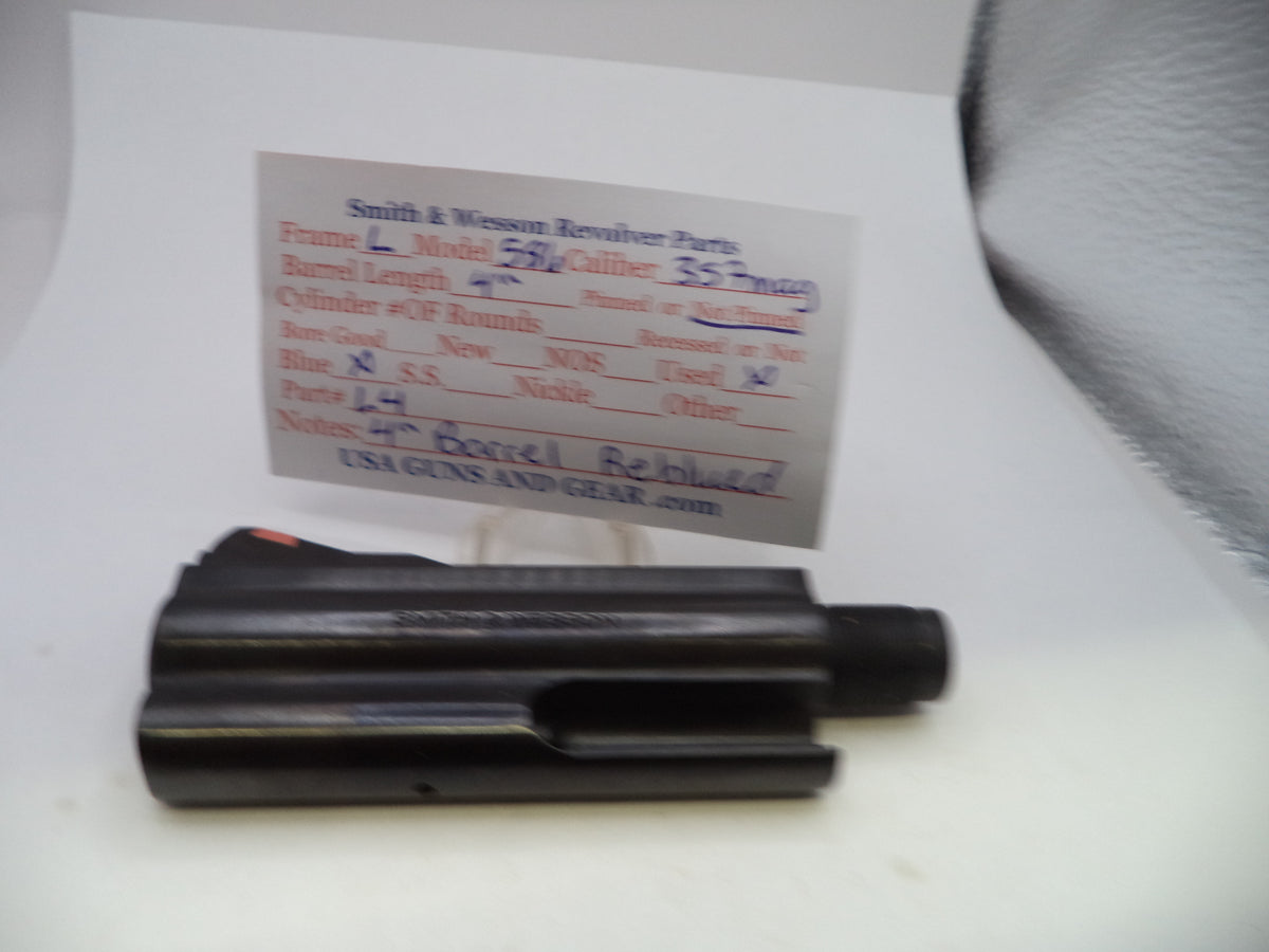 A black metal revolver part, likely a frame or cylinder component, displayed on a white surface with a handwritten parts label card behind it.