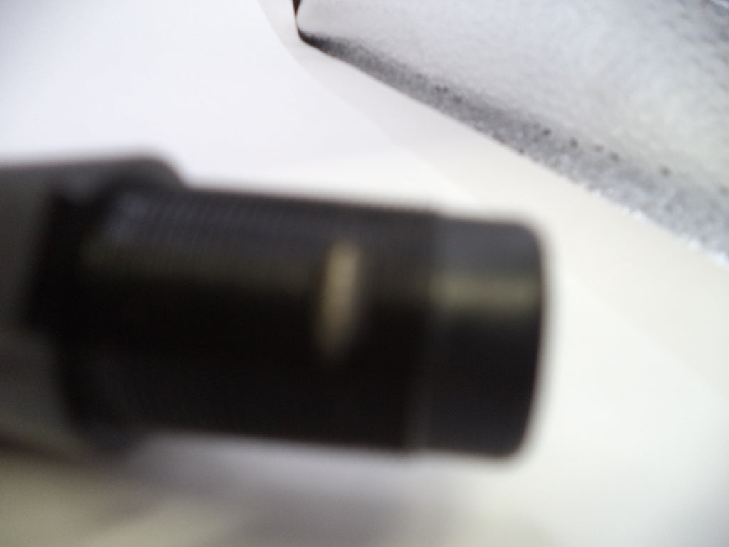 A close-up, heavily blurred shot of a black cylindrical object on a white surface, with part of a textured gray item visible in the upper right.