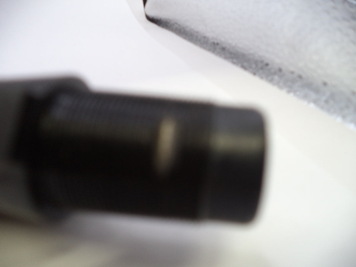 A close-up, heavily blurred shot of a black cylindrical object on a white surface, with part of a textured gray item visible in the upper right.