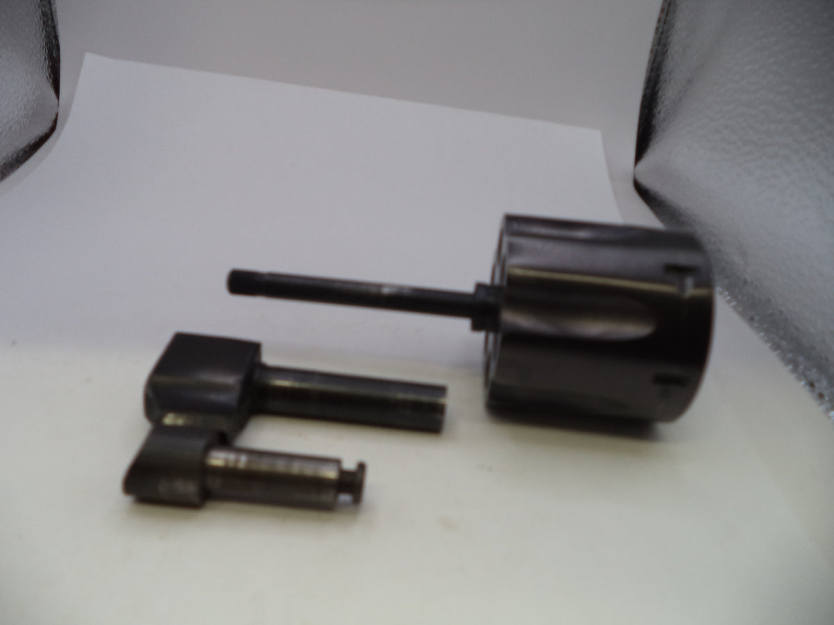 A set of dark metal mechanical parts, including a cylindrical component with a protruding rod and two small square-shaped metal pieces, displayed on a white surface.