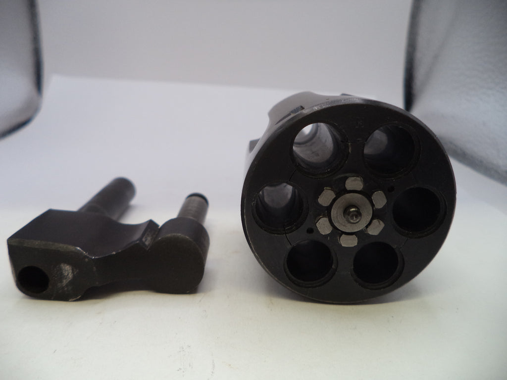 A close-up of a black multi-shot revolver cylinder and a separate black metal component on a white surface.