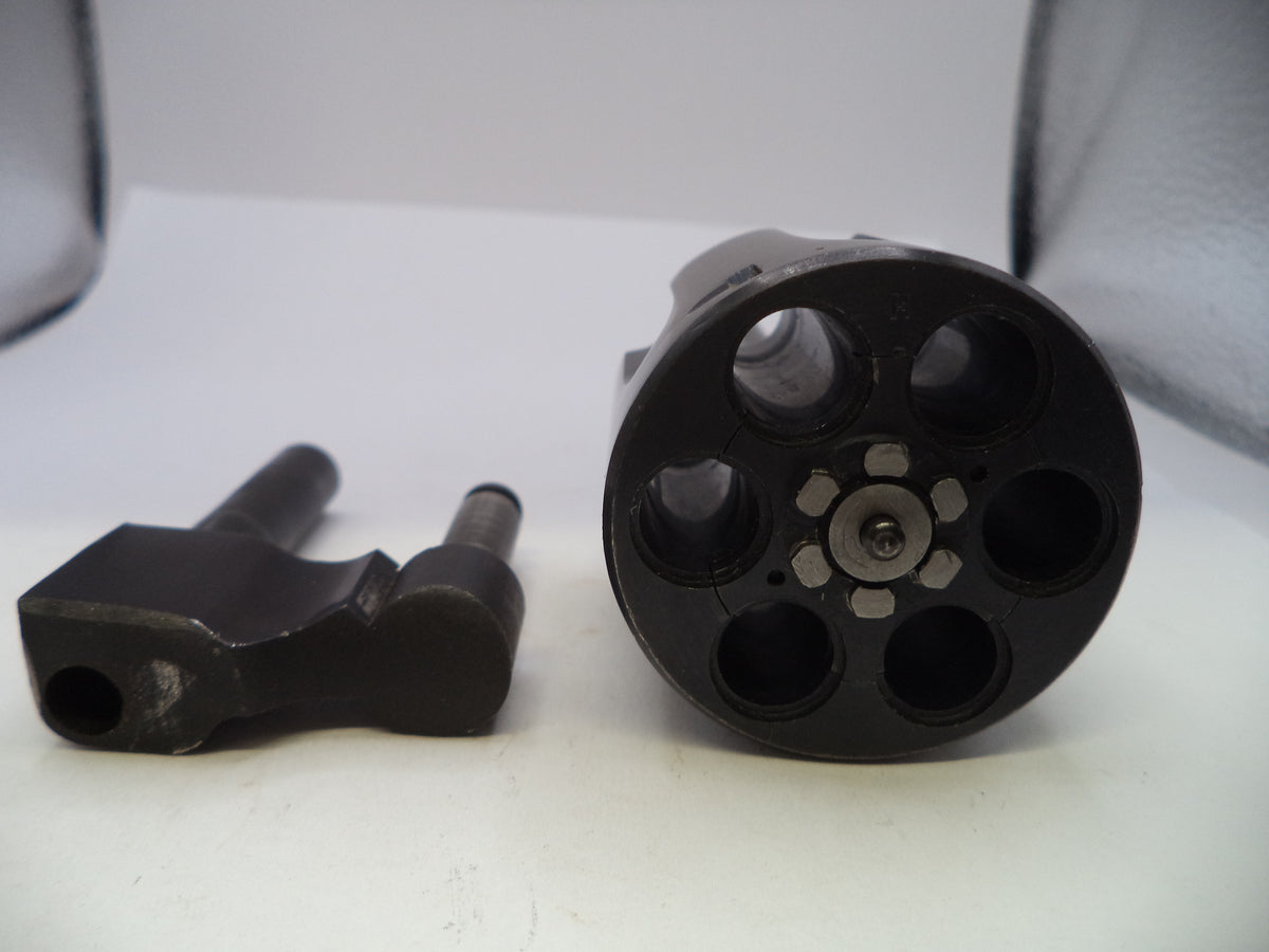 A close-up of a black multi-shot revolver cylinder and a separate black metal component on a white surface.