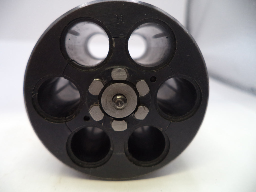 A close-up of a black circular mechanical component with six circular openings and a central metallic screw fastener, photographed on a white background.