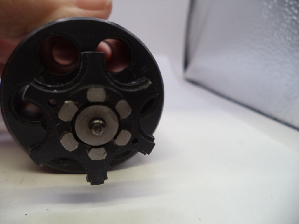 A close-up of a black revolver cylinder end with a metallic center pin and six circular cutouts, held by a hand against a white background with a silver textured object in the upper right.