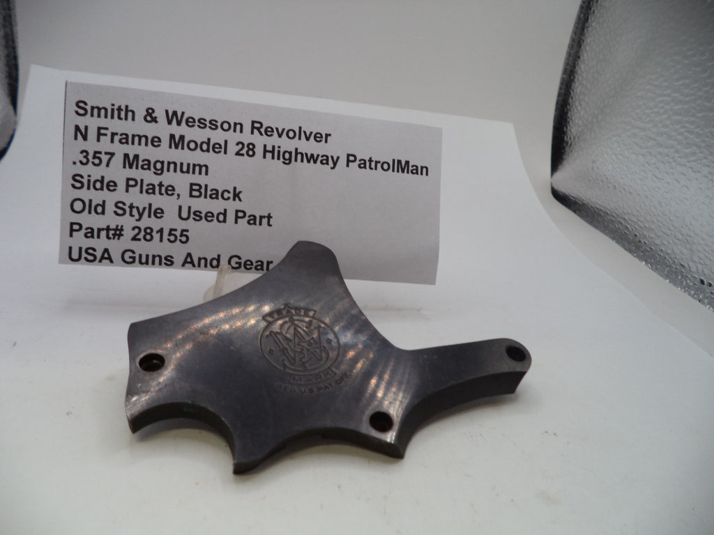 A black metal side plate part for a Smith & Wesson revolver, displayed on a white surface with a paper label card behind it.