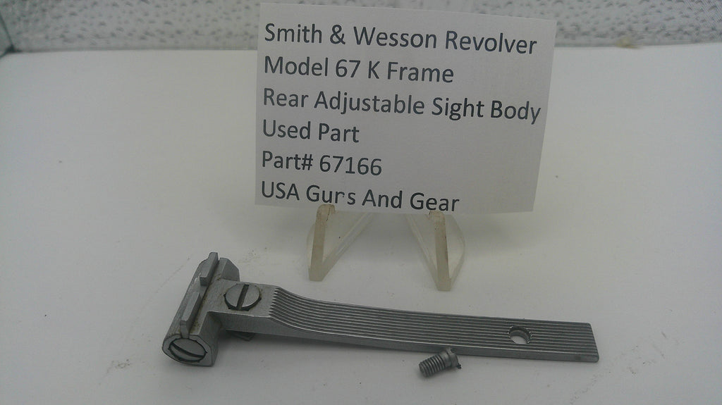A metal Smith & Wesson revolver sight body part for Model 67 K, displayed on a white surface with a small white label card behind it that reads 'Smith & Wesson Revolver Model 67 K Frame Rear Adjustable Sight Body Used Part Part# 67166 USA Gurs And Gear'.