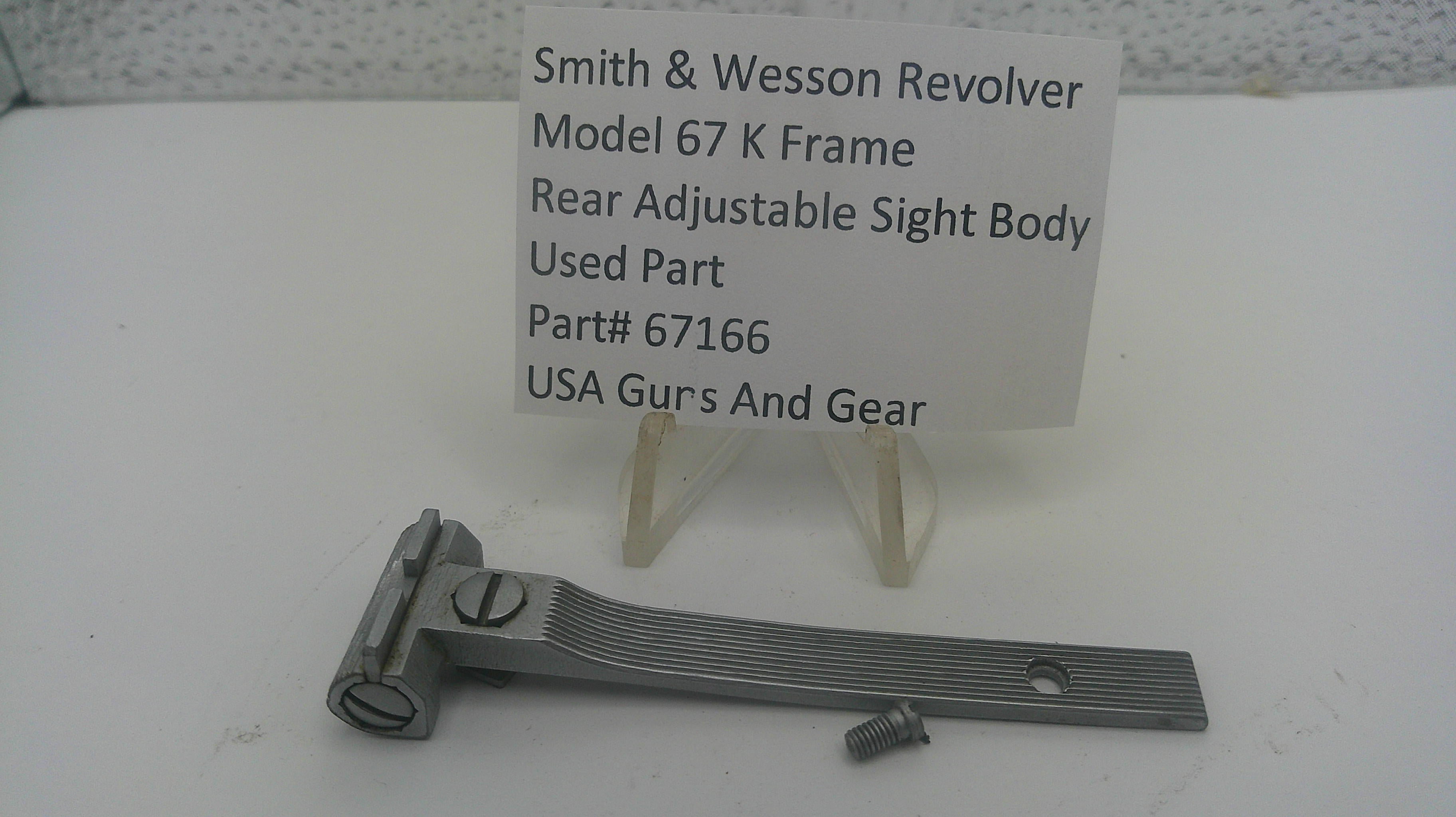 A metal Smith & Wesson revolver sight body part for Model 67 K, displayed on a white surface with a small white label card behind it that reads 'Smith & Wesson Revolver Model 67 K Frame Rear Adjustable Sight Body Used Part Part# 67166 USA Gurs And Gear'.