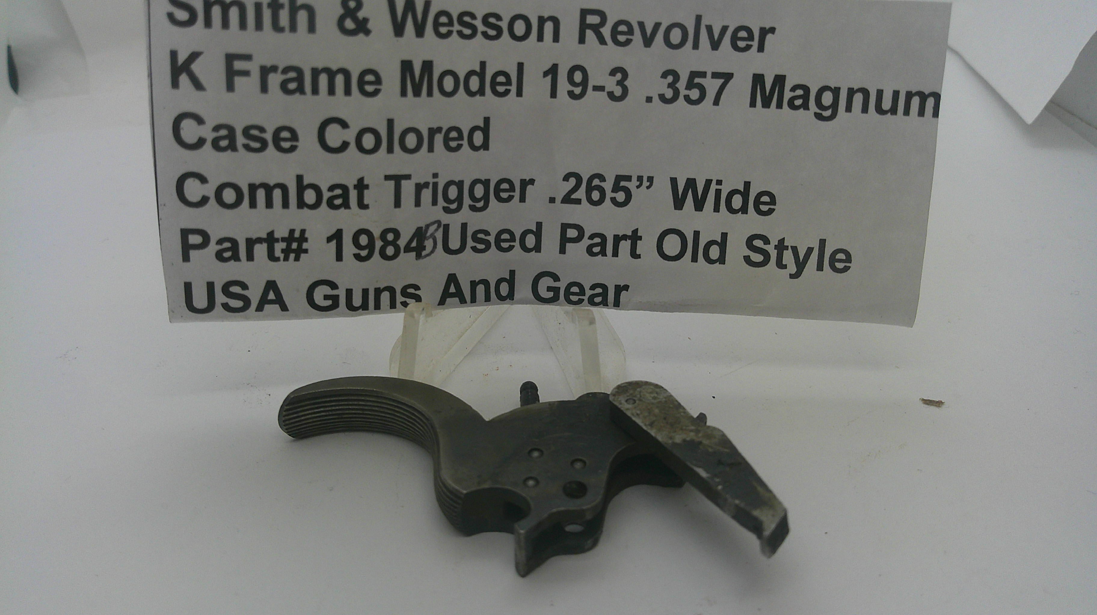 A used metal Smith & Wesson revolver trigger part, model 19-3 .357 Magnum, shown on a white surface with a handwritten paper label in the background.