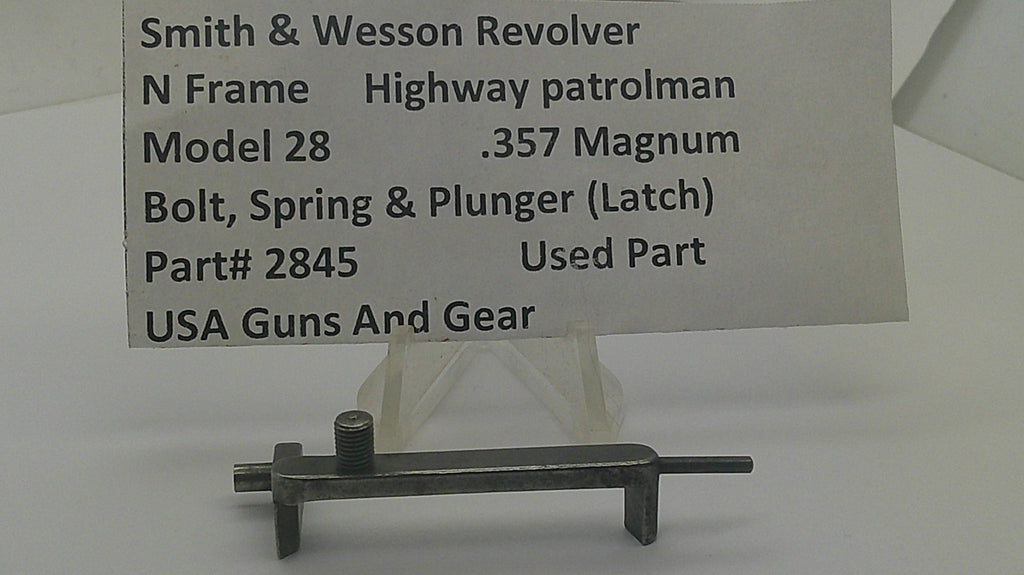 A used metal latch part for a Smith & Wesson revolver, displayed on a clear stand with a paper label card in the background. The label details the part number 2845 and notes it is a used part for a Model 28 .